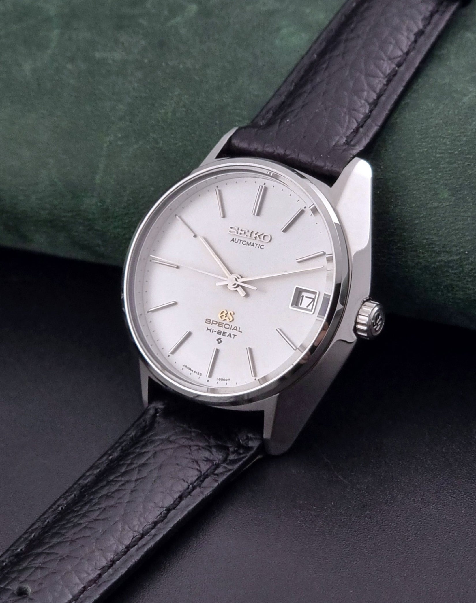 Vintage Grand Seiko Special 6155-8000 Silver wristwatch with black leather strap on gray background crown view