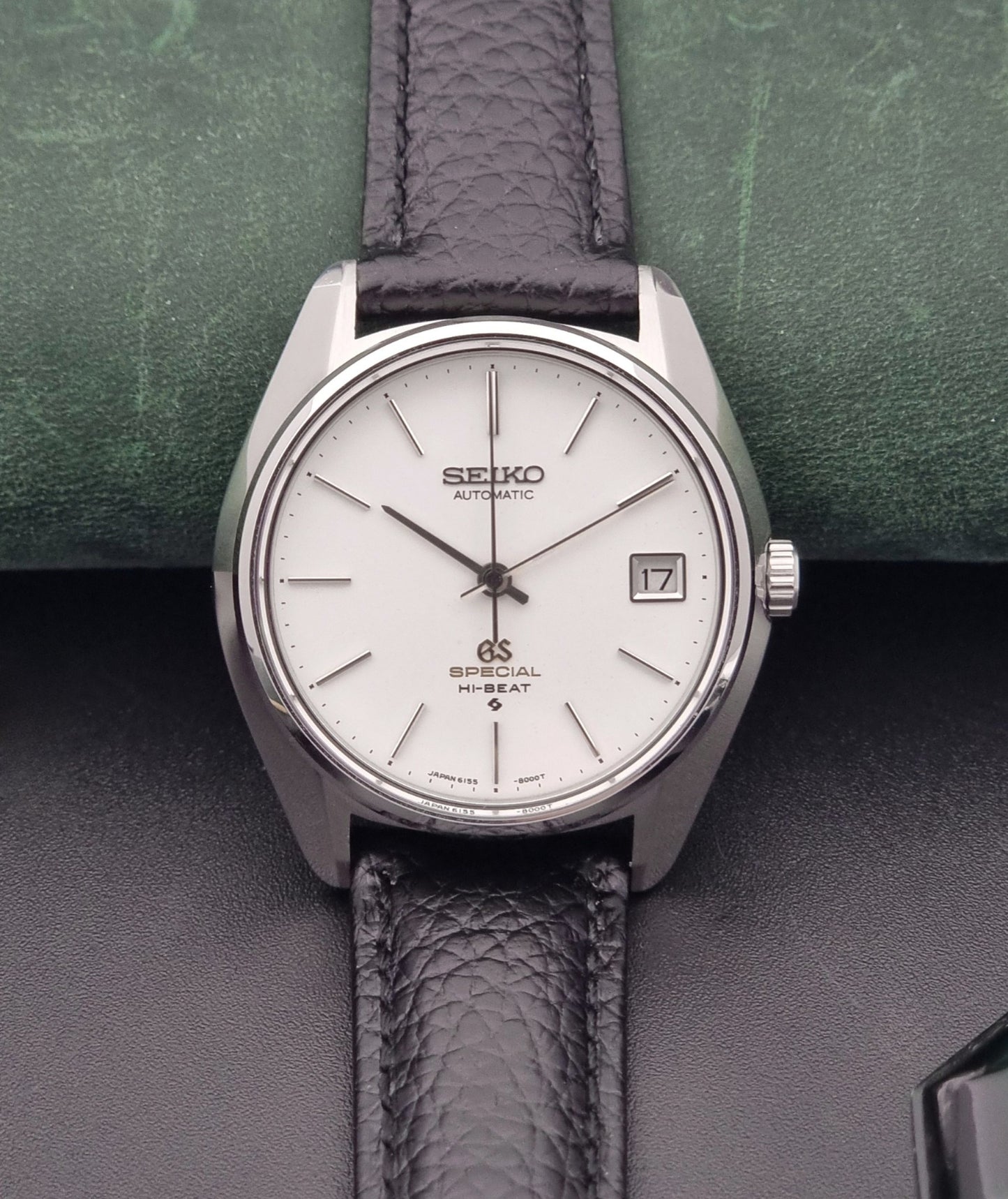 Vintage Grand Seiko Special 6155-8000 Silver wristwatch with black leather strap on a green and gray background close up dial view