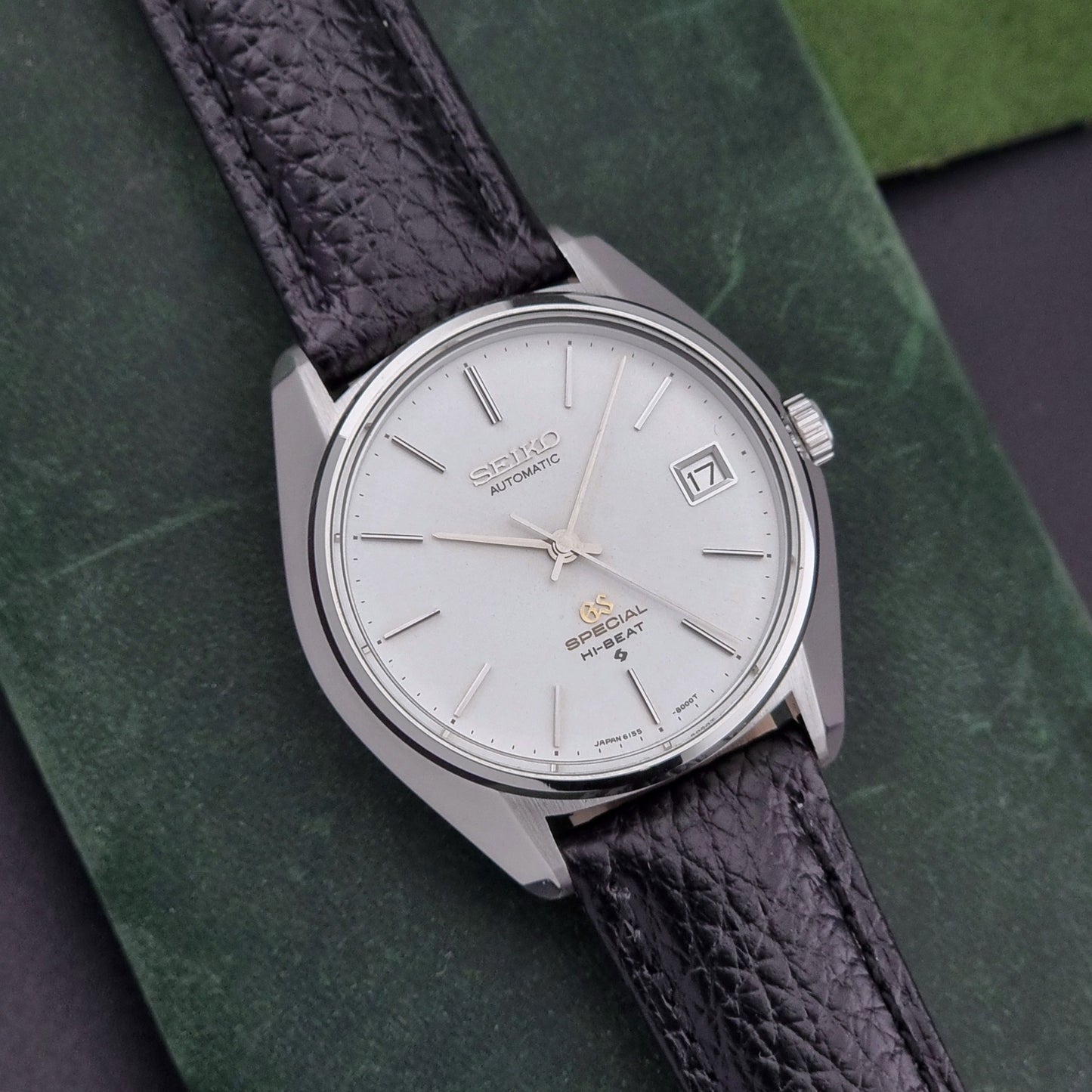 Vintage Grand Seiko Special 6155-8000 Silver wristwatch with black leather strap on a green and gray background close dial view