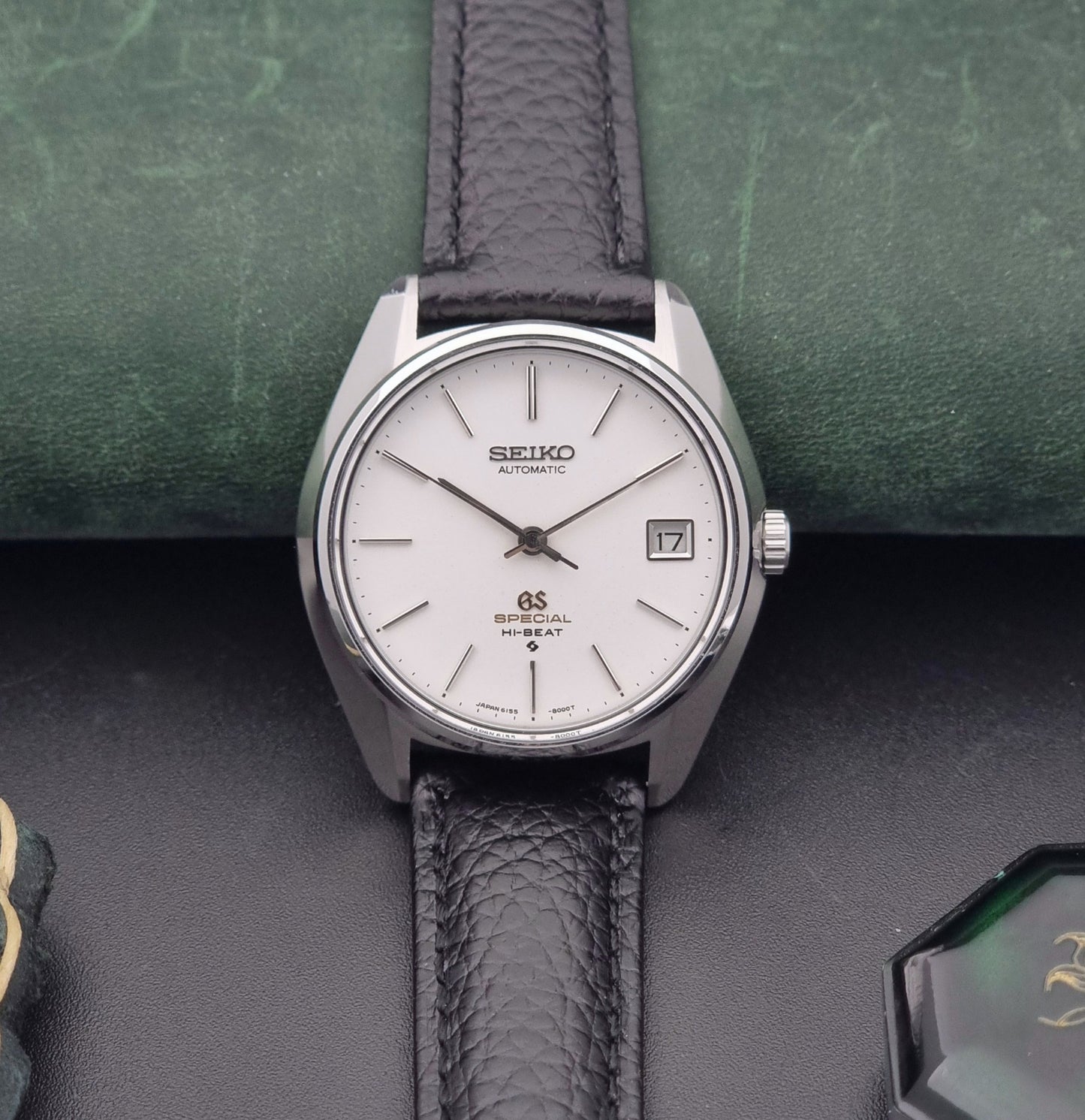Vintage Grand Seiko Special 6155-8000 Silver wristwatch with black leather strap on a green and gray background front view