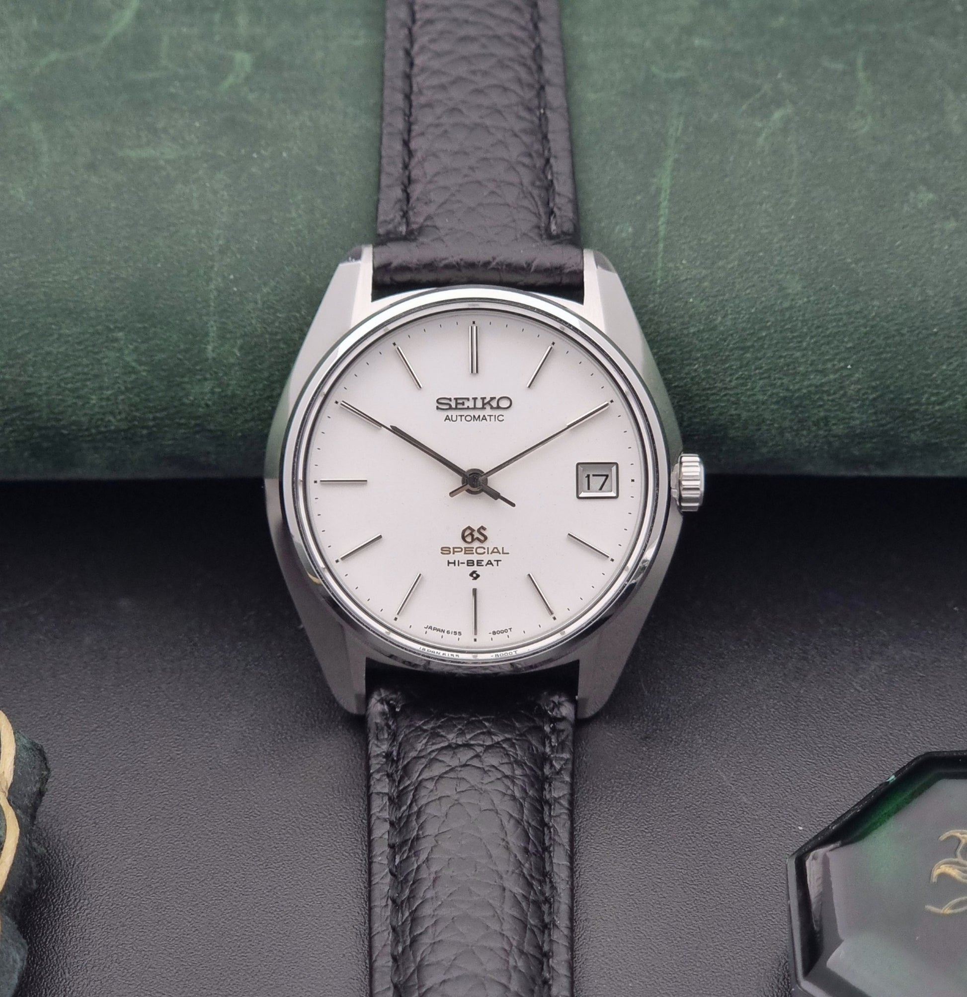 Vintage Grand Seiko Special 6155-8000 Silver wristwatch with black leather strap on a green and gray background front view