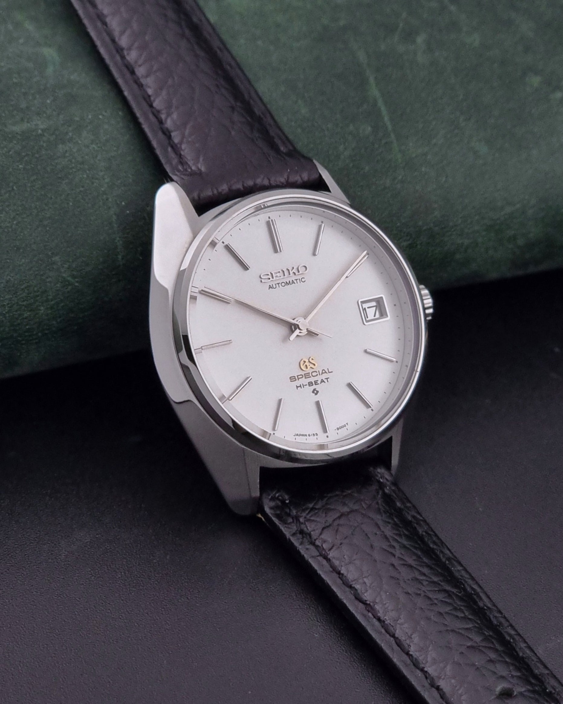 Vintage Grand Seiko Special 6155-8000 Silver wristwatch with black leather strap on a green and gray background side view