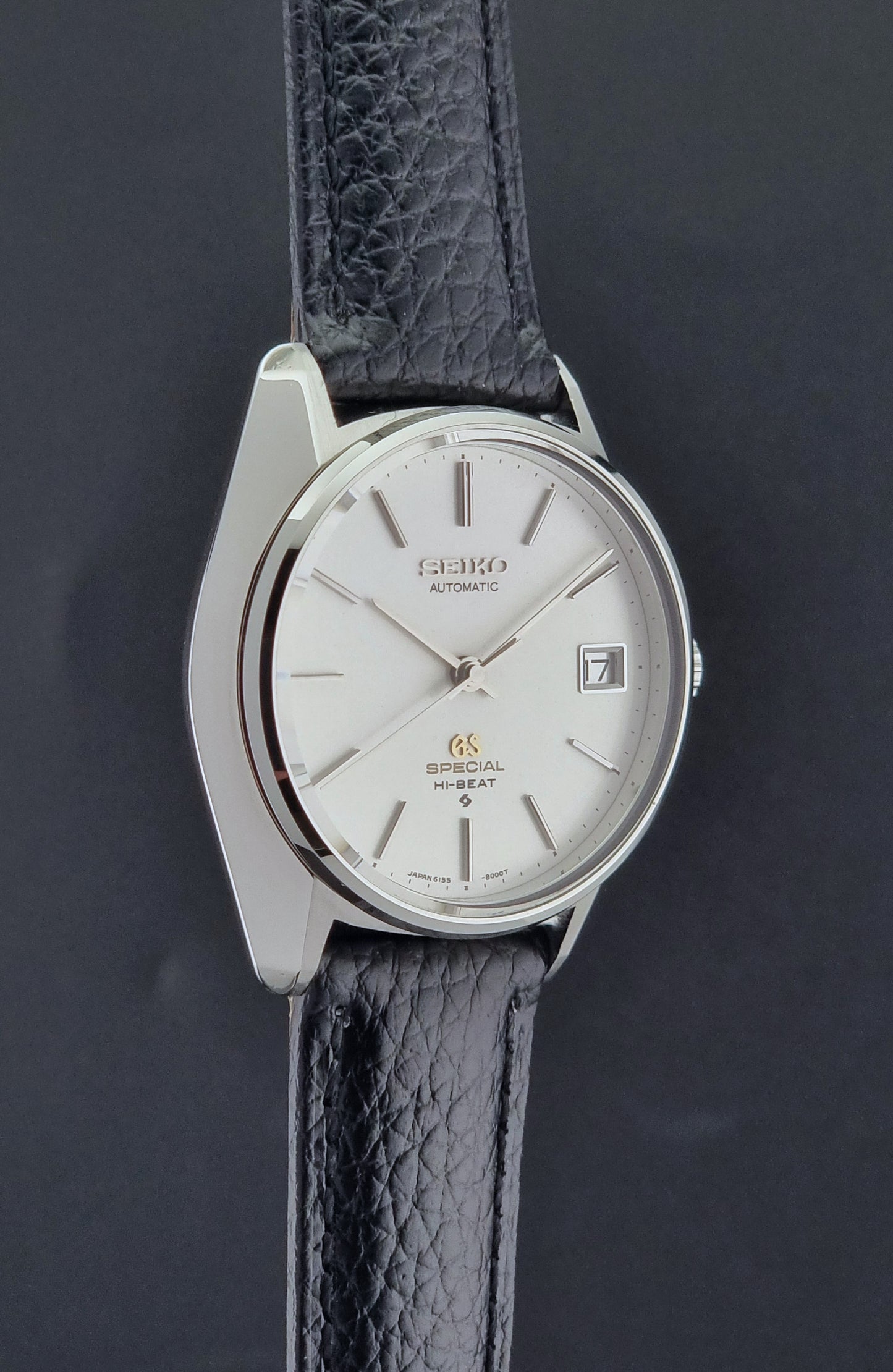 Vintage Grand Seiko Special 6155-8000 Silver wristwatch with black leather strap on a gray background side profile view