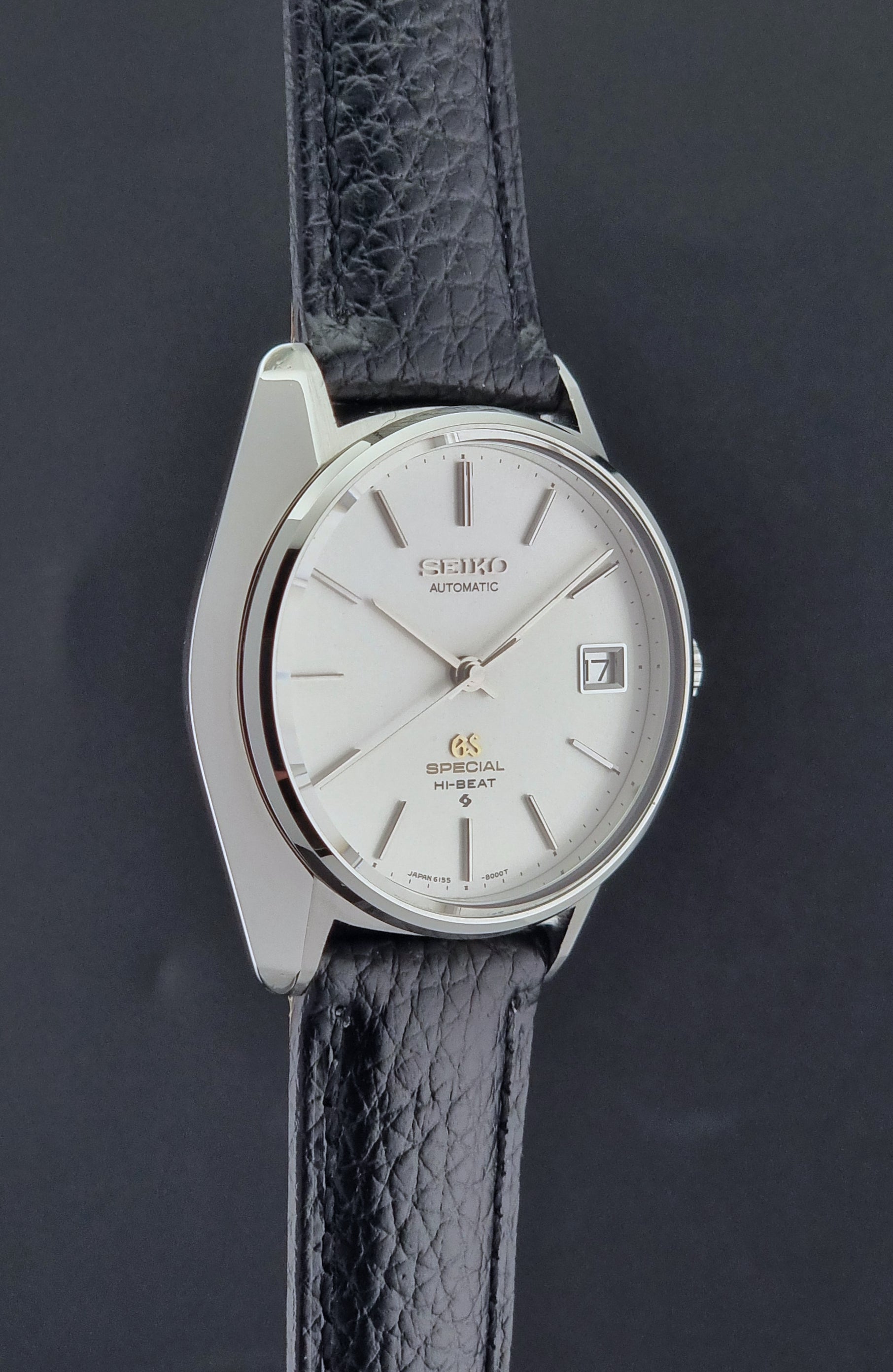 Vintage Grand Seiko Special 6155-8000 Silver wristwatch with black leather strap on a gray background side profile view
