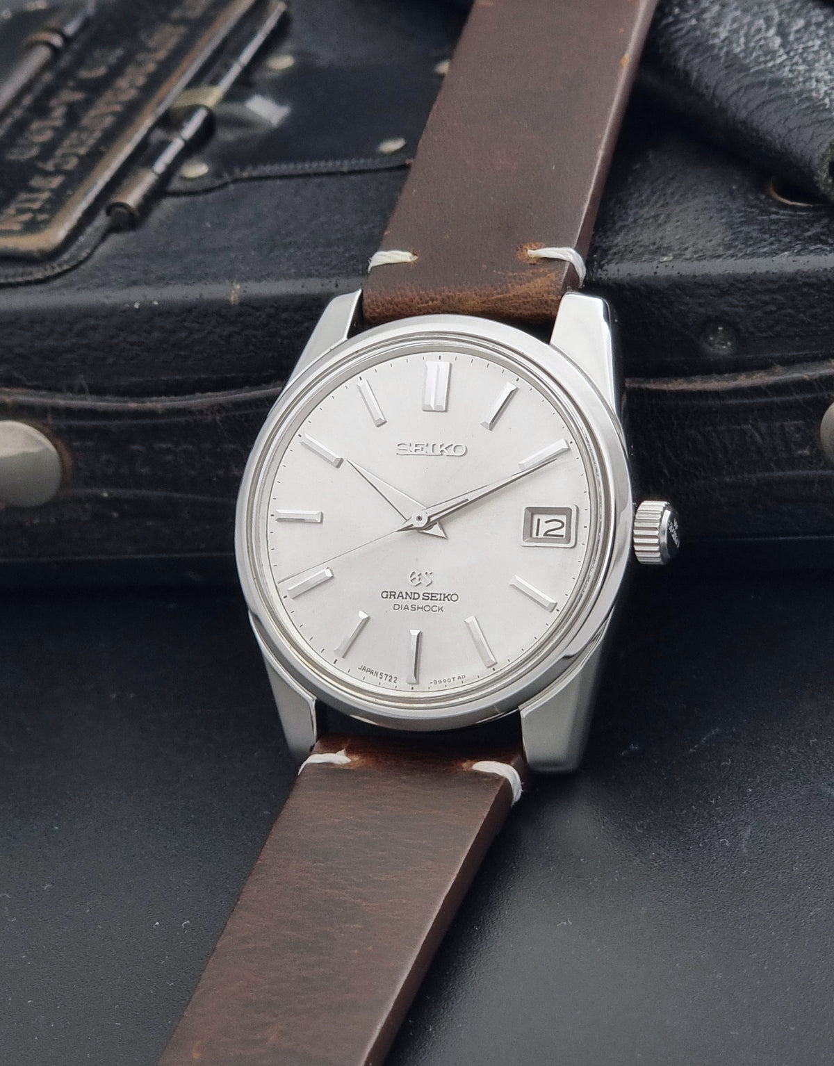 Vintage Grand Seiko 5722-9991 wristwatch with a silver dial, silver case, and a brown leather strap, shown on a dark gray background crown view
