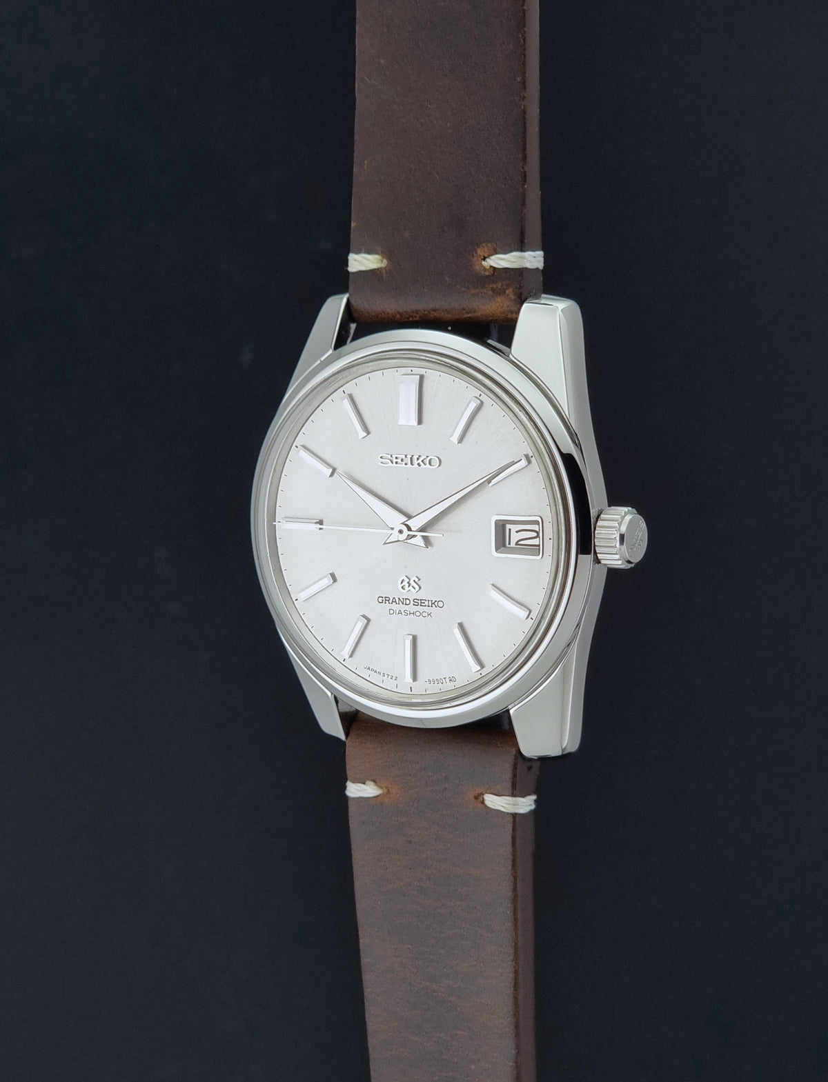 Vintage Grand Seiko 5722-9991 wristwatch with a silver dial, silver case, and a brown leather strap, shown on a dark gray background crown profile view