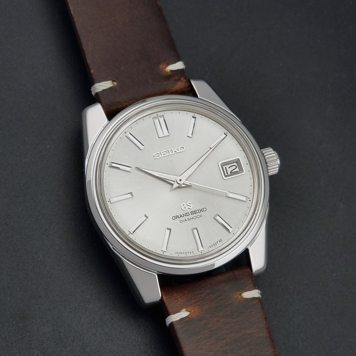 Vintage Grand Seiko 5722-9991 wristwatch with a silver dial, silver case, and a brown leather strap, shown on a dark gray background front view