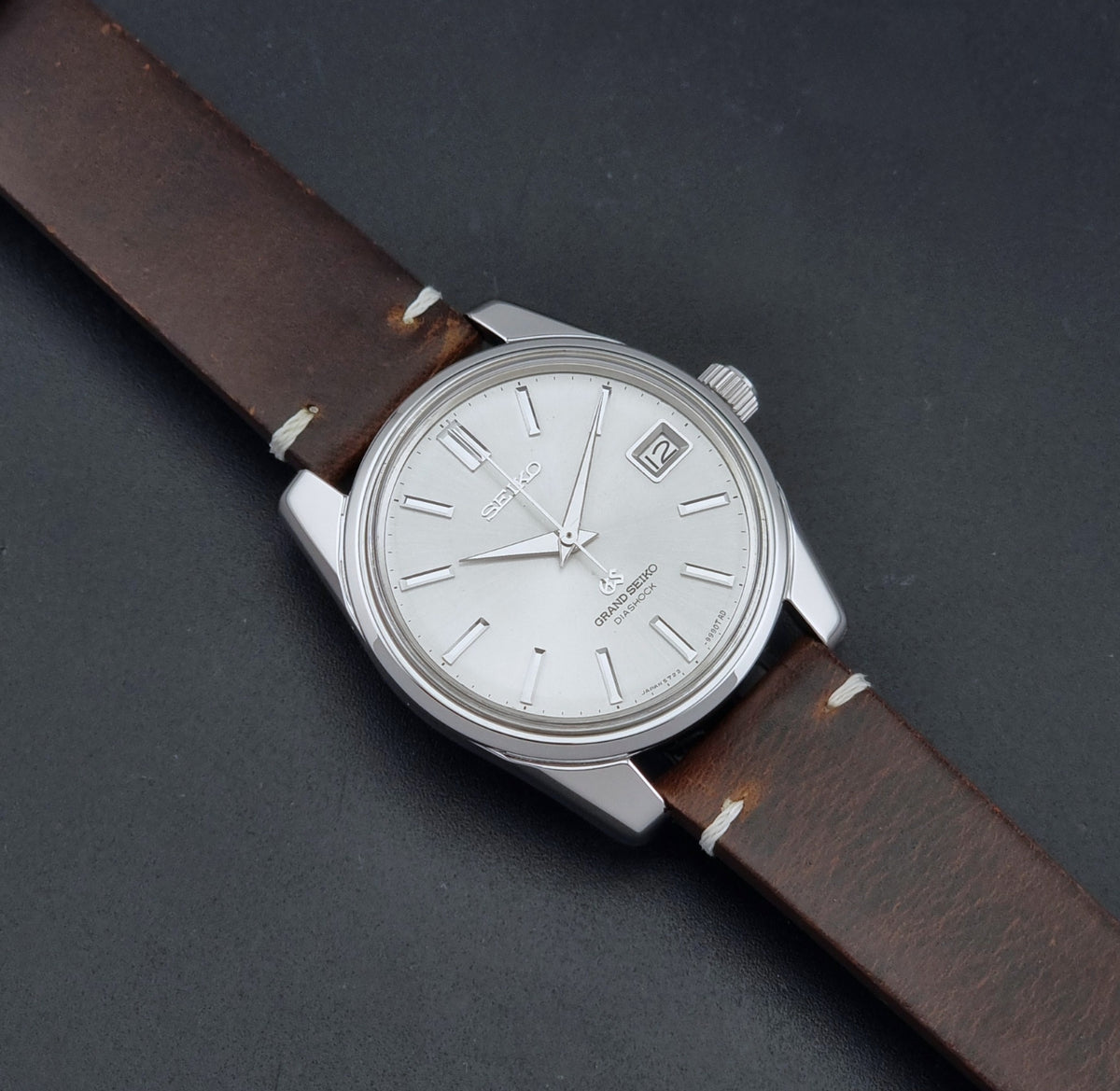 Vintage Grand Seiko 5722-9991 wristwatch with a silver dial, silver case, and a brown leather strap, shown on a dark gray background dial