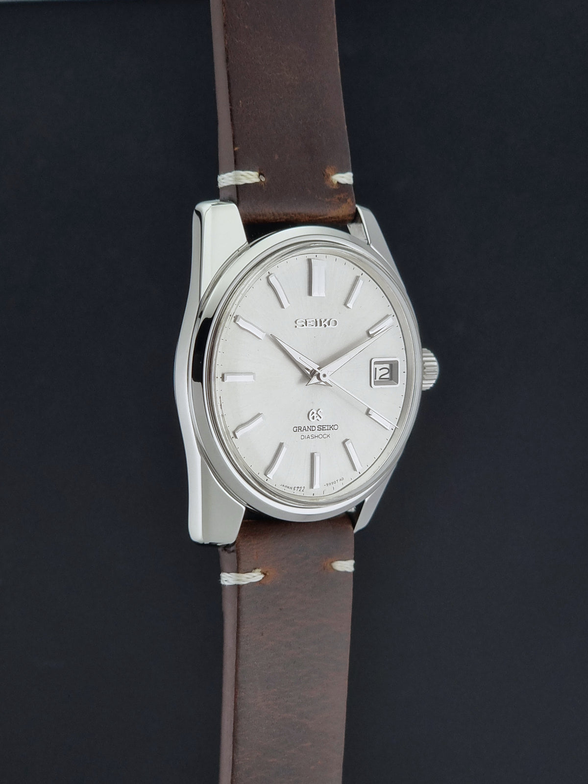 Vintage Grand Seiko 5722-9991 wristwatch with a silver dial, silver case, and a brown leather strap, shown on a dark gray background side profile view