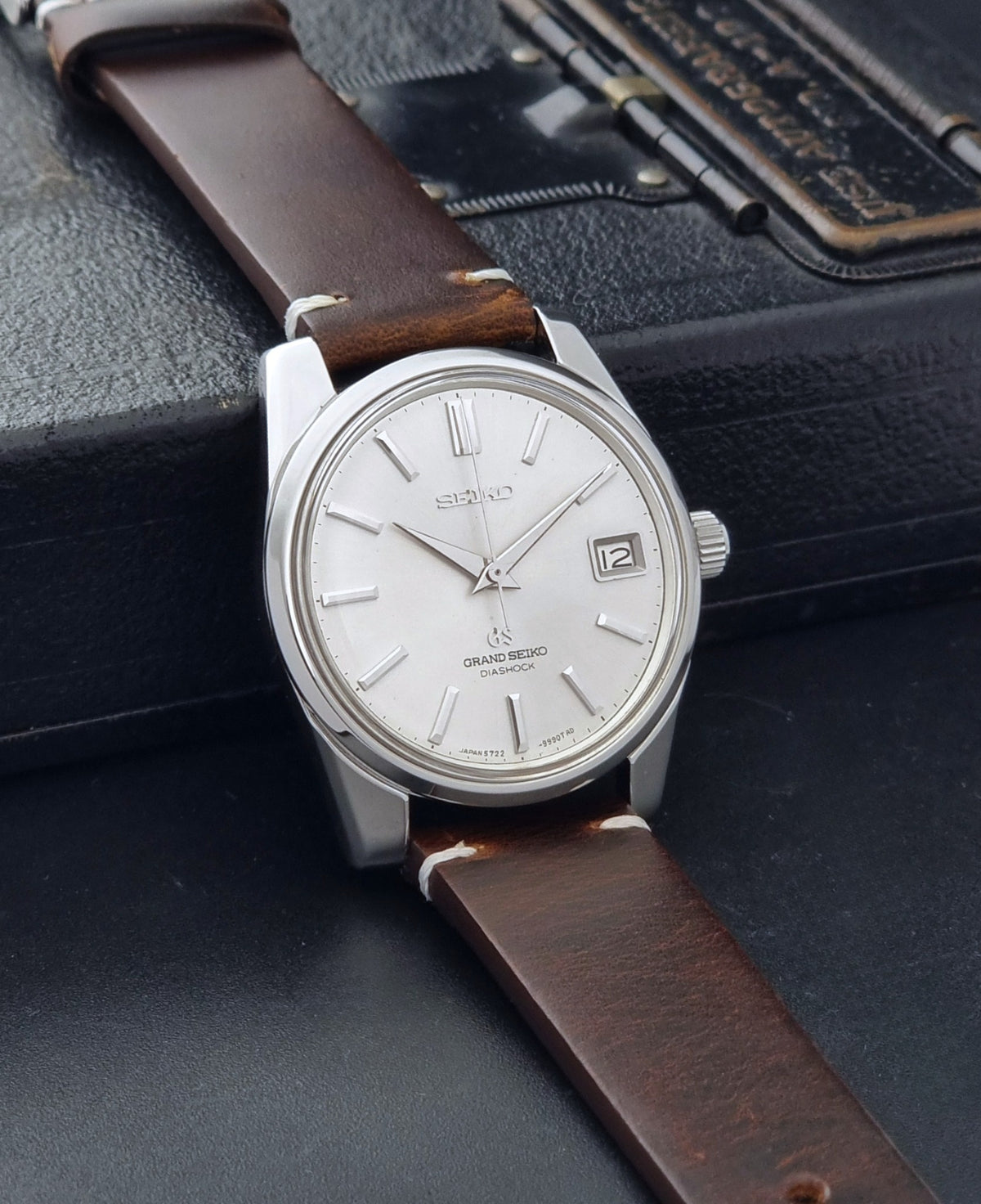 Vintage Grand Seiko 5722-9991 wristwatch with a silver dial, silver case, and a brown leather strap, shown on a dark gray background side view