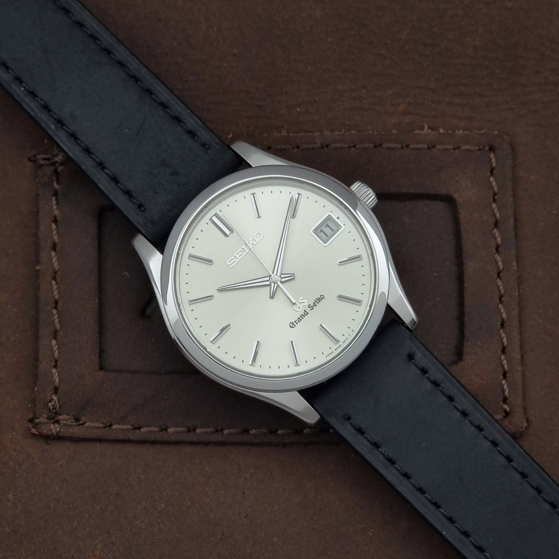 Grand Seiko SBGV001 quartz watch 9F82-0A10 with a silver-toned case,  and black leather strap, on a brown background dial