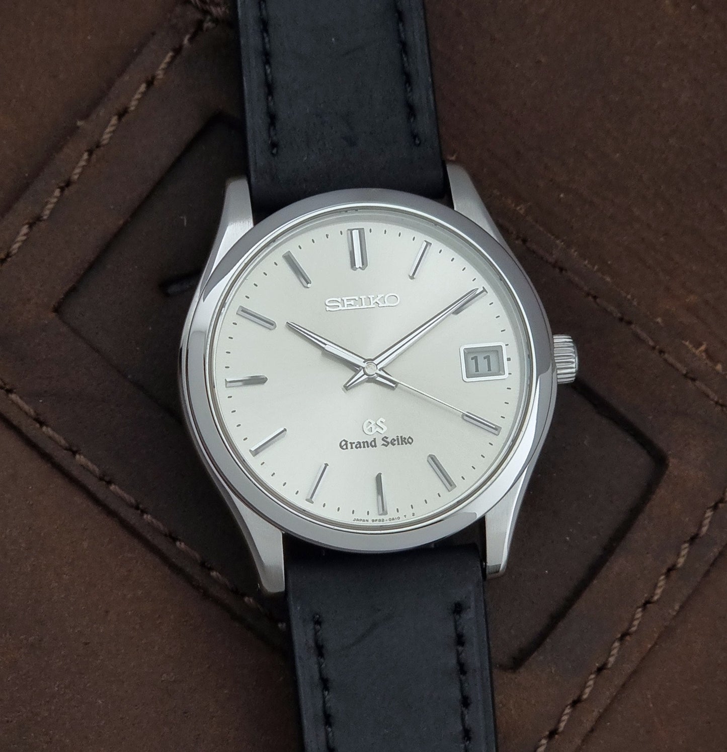 Grand Seiko SBGV001 quartz watch 9F82-0A10 with a silver-toned case,  and black leather strap, on a brown background front view