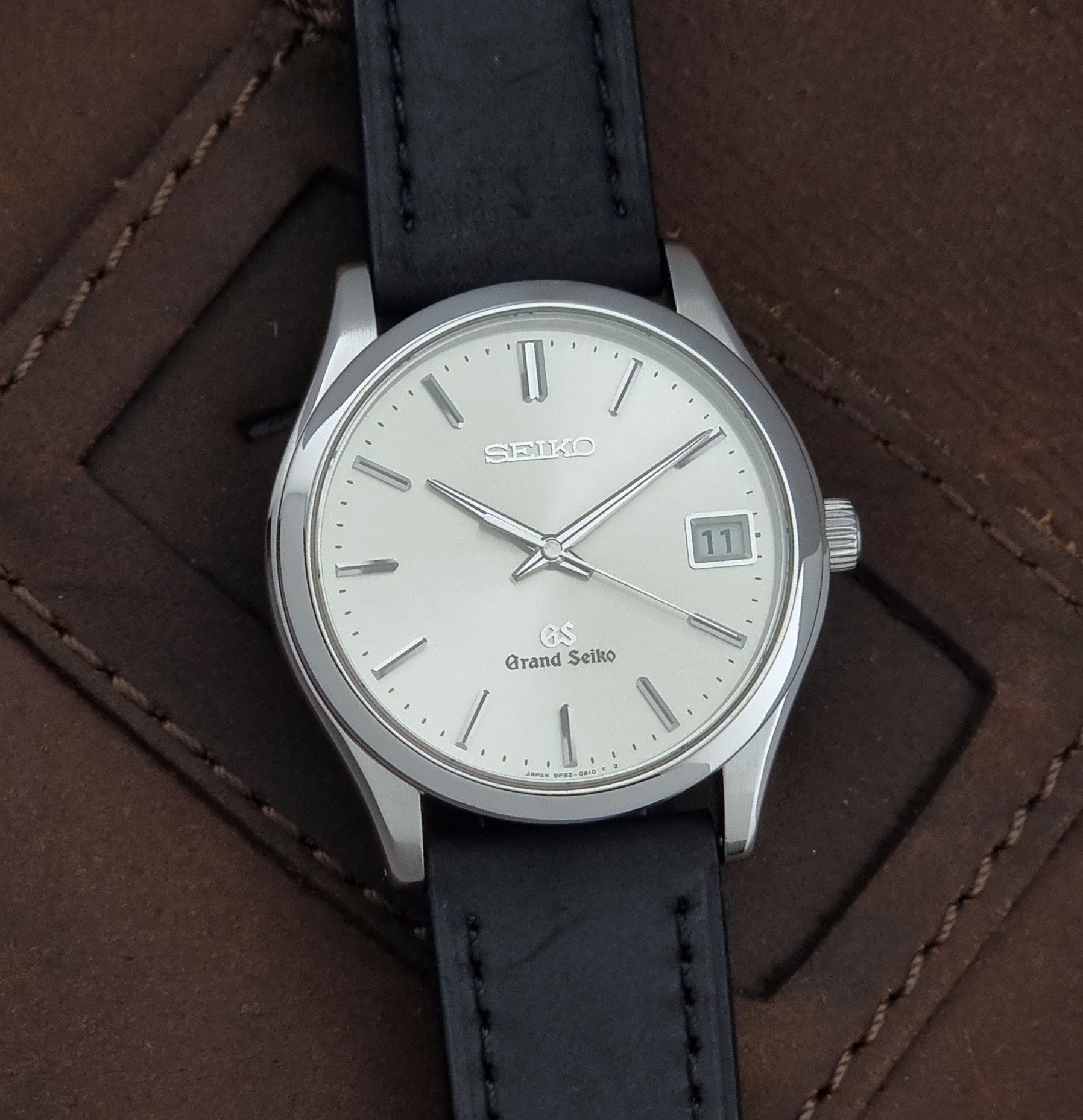 Grand Seiko SBGV001 quartz watch 9F82-0A10 with a silver-toned case,  and black leather strap, on a brown background front view