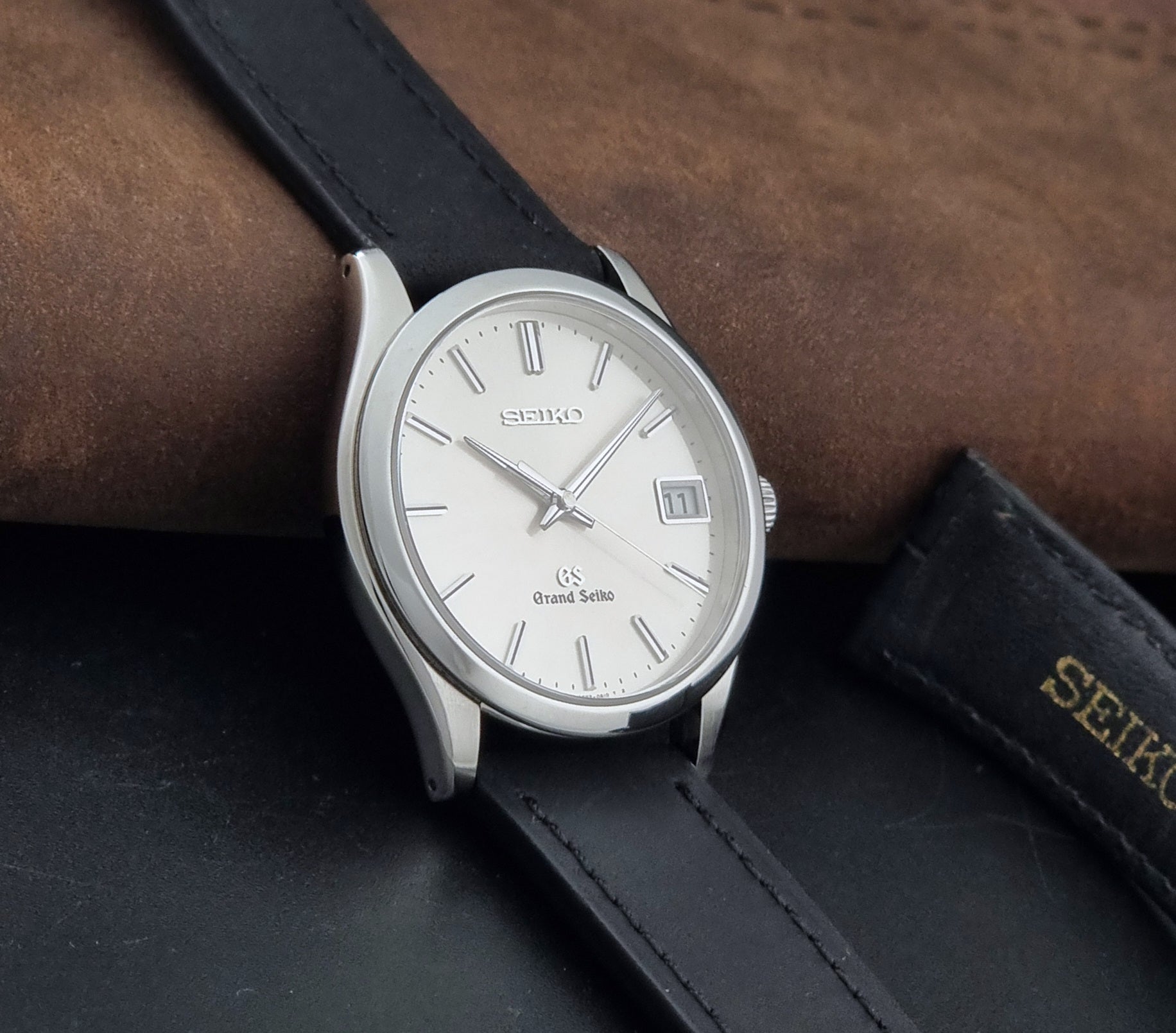 Grand Seiko SBGV001 quartz watch 9F82-0A10 with a silver-toned case,  and black leather strap, on a brown background side view
