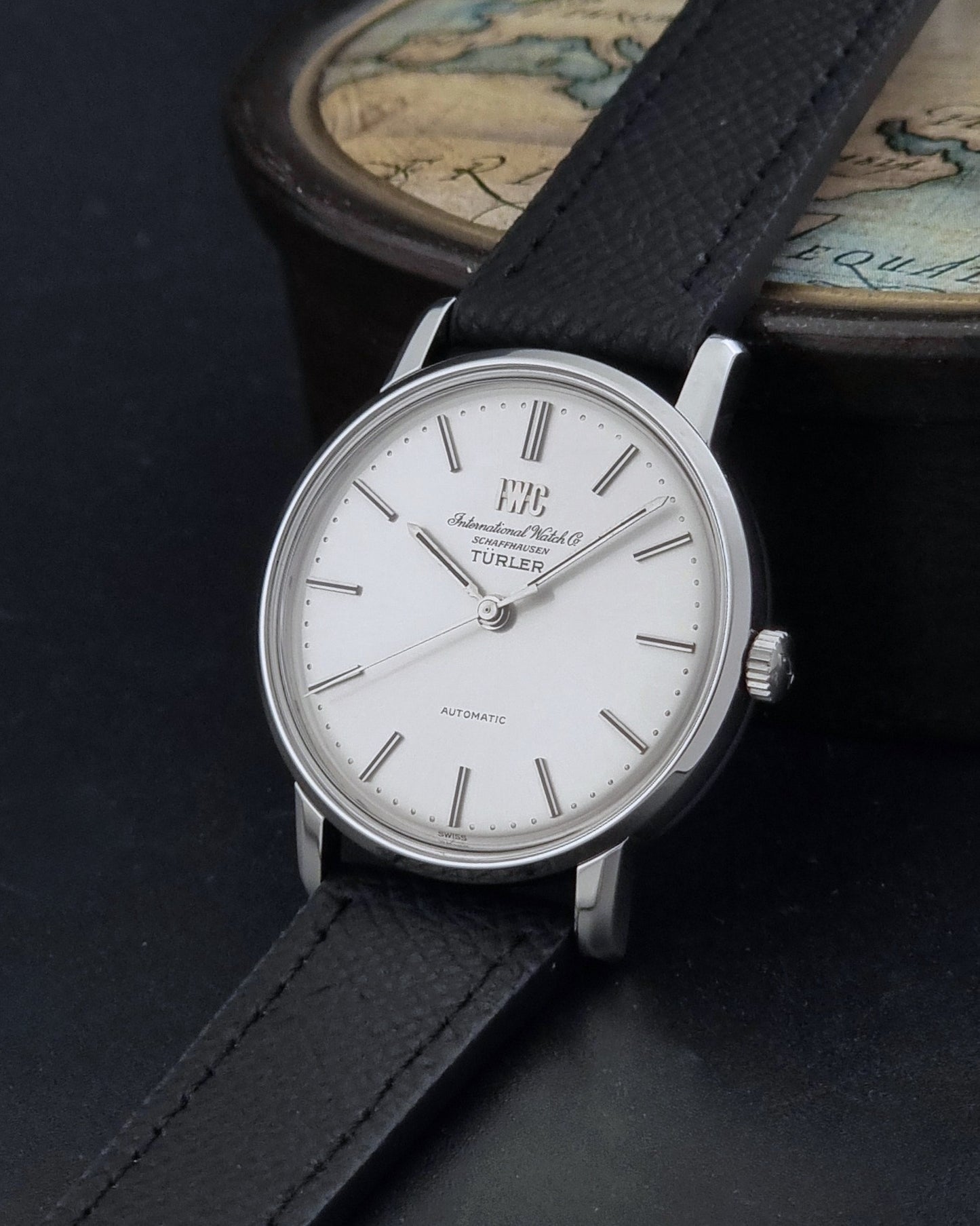 Vintage IWC R802A watch with a silver-toned case,  and textured black leather strap, on a dark background crown view