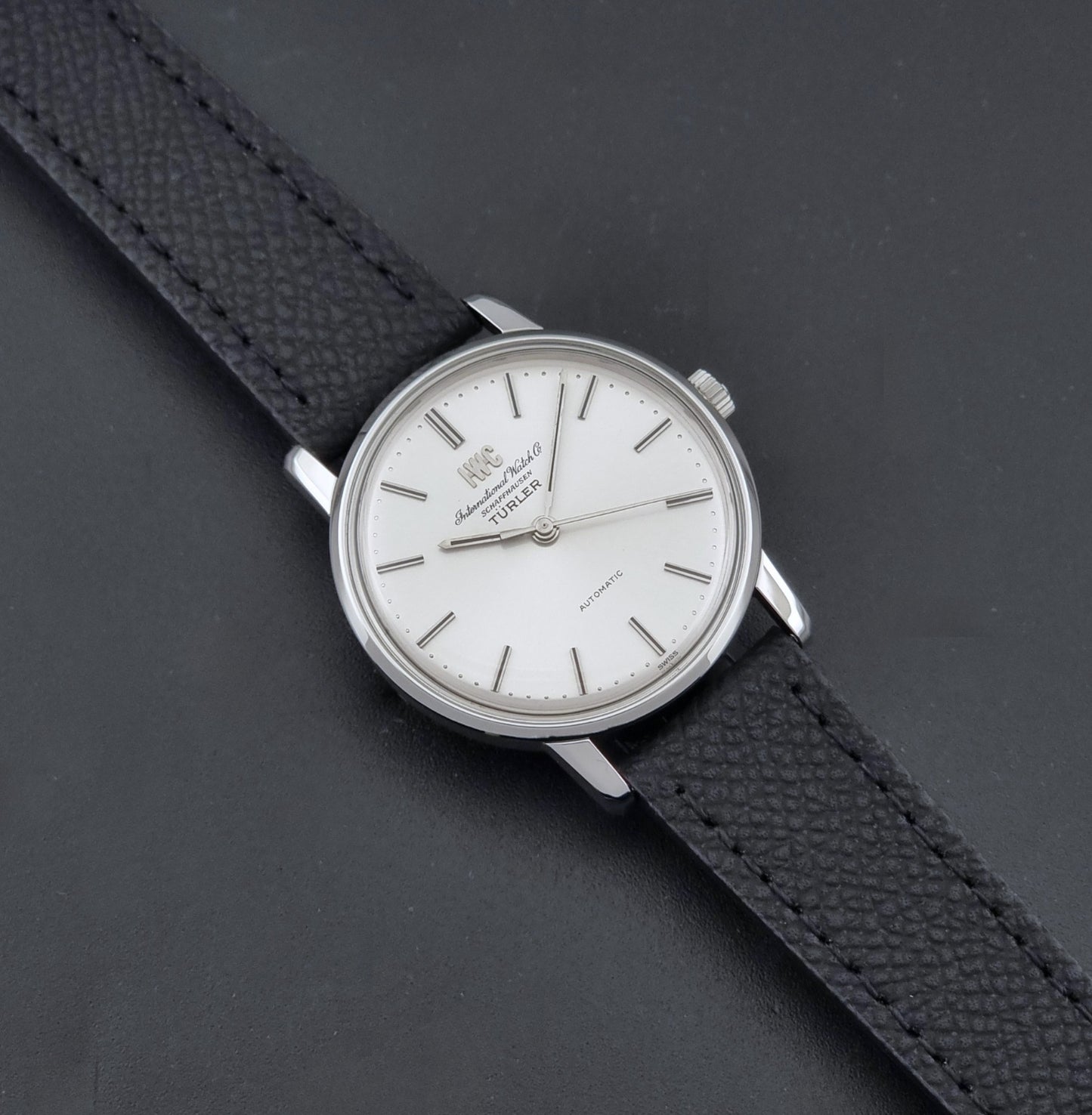 Vintage IWC R802A watch with a silver-toned case,  and textured black leather strap, on a dark background dial