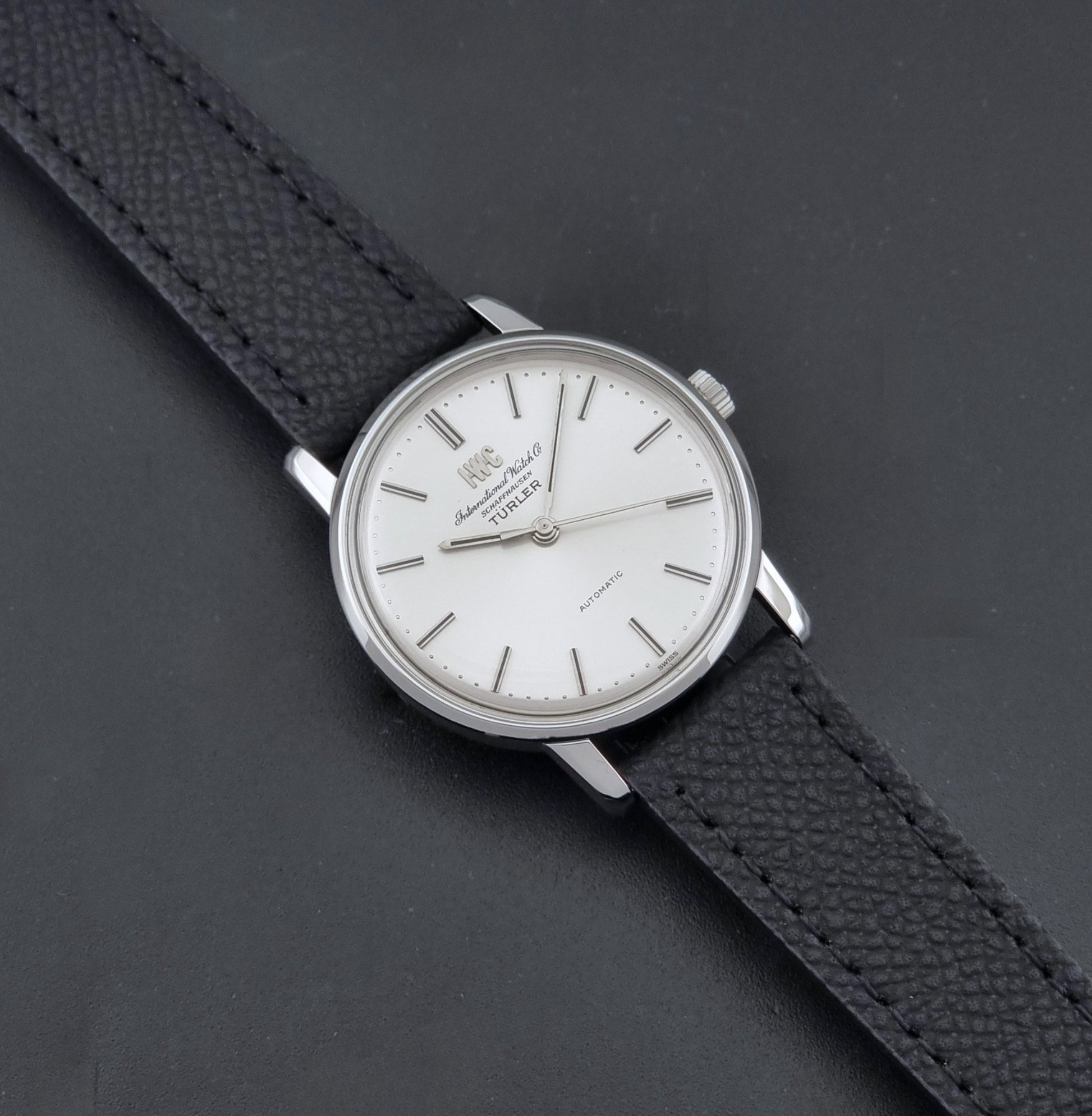 Vintage IWC R802A watch with a silver-toned case,  and textured black leather strap, on a dark background dial