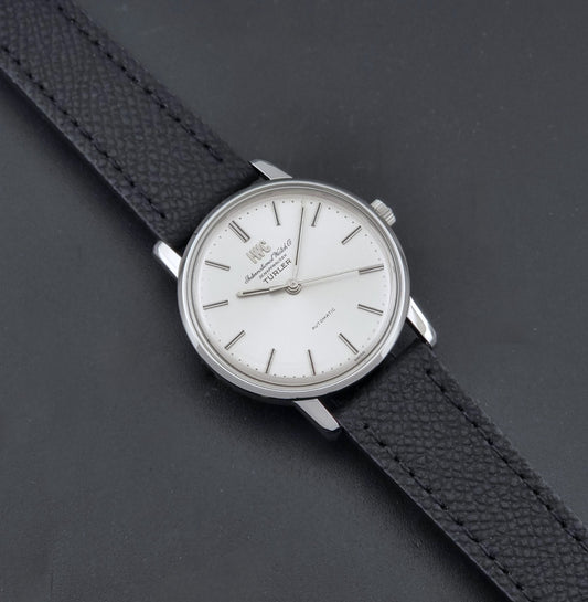 Vintage IWC R802A watch with a silver-toned case,  and textured black leather strap, on a dark background dial