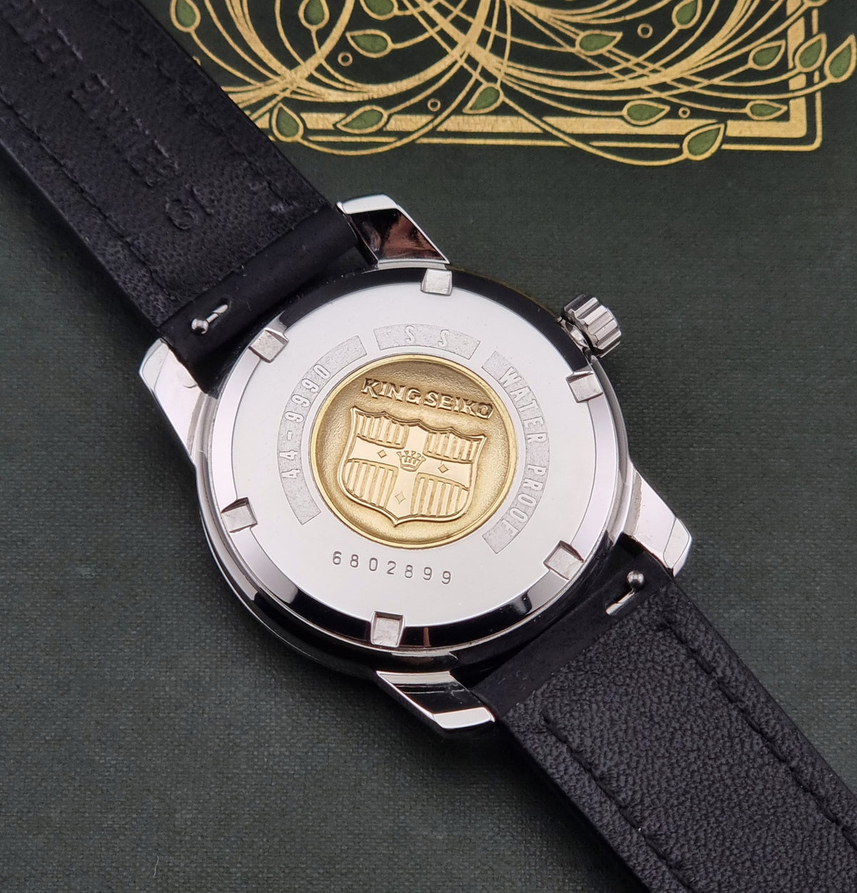 Close-up of the back of a wristwatch with a black leather strap, silver-tone case, and a prominent gold-tone crest emblem on the case back. The watch is photographed on a dark green textured surface with a gold decorative pattern in the background.