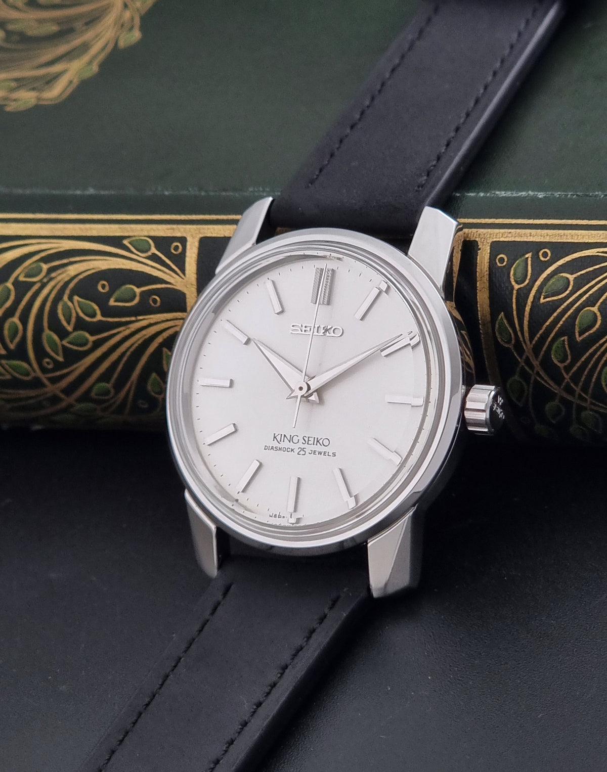 A Seiko King Seiko wristwatch with a silver-toned oval case, white dial, and black leather strap, styled on a dark surface with an ornate green book in the background.
