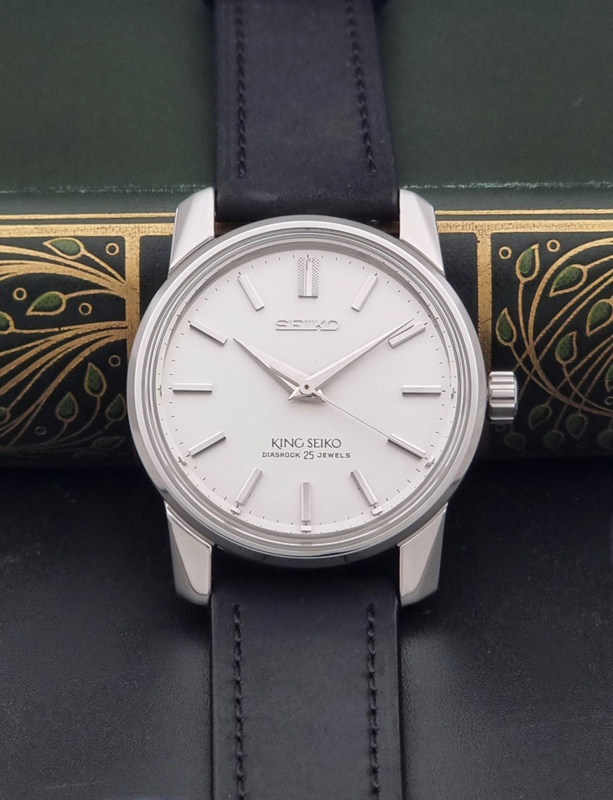 Vintage King Seiko watch 44-9990 with a silver-toned stainless steel case, white dial, and black leather strap, displayed on a dark textured surface with a decorative book in the background.