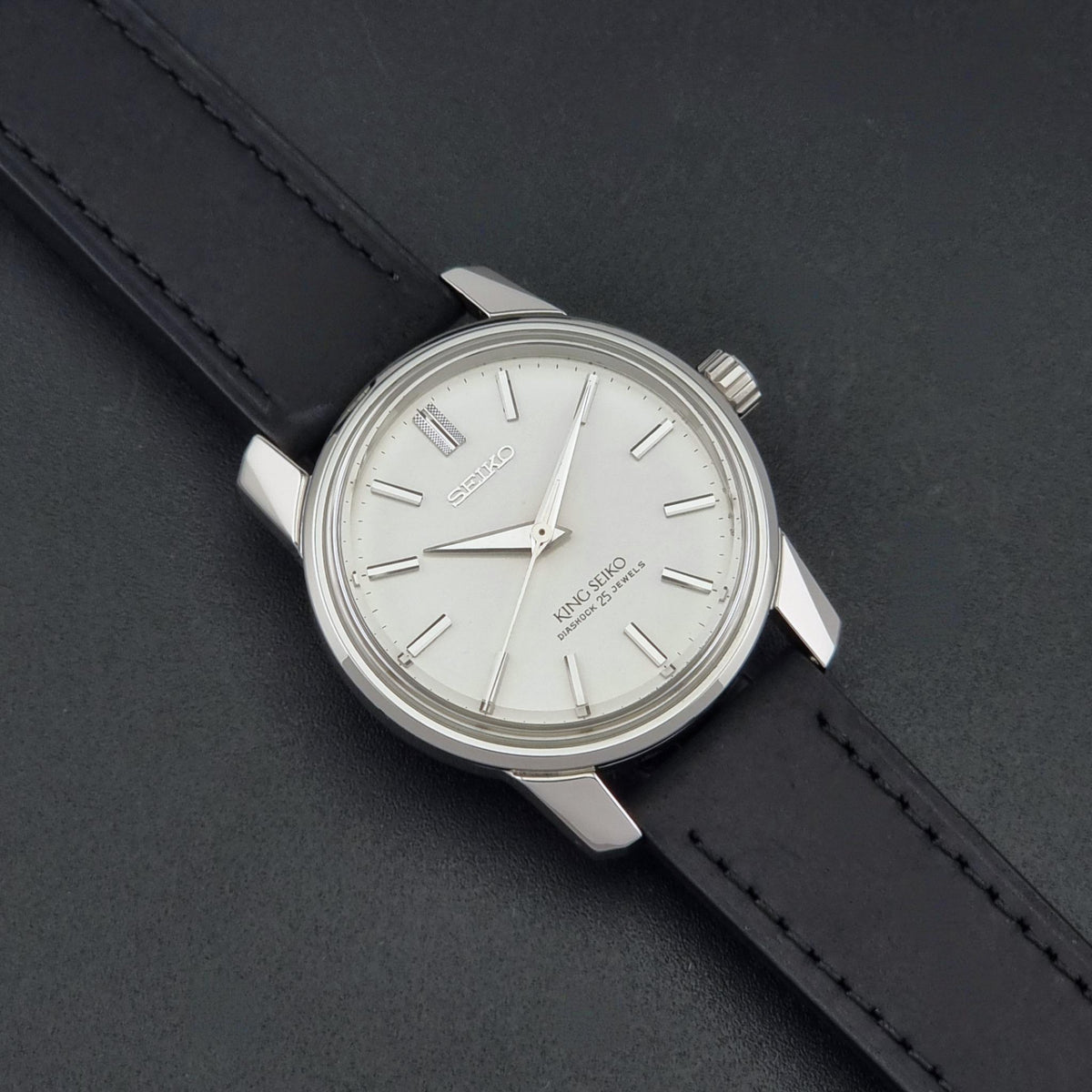 Vintage King Seiko watch 44-9990 with a silver-toned metal case, white dial, and black leather strap, photographed diagonally on a dark textured background.