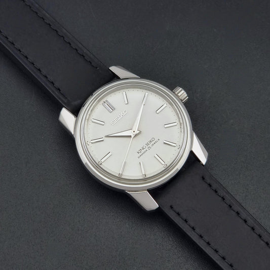 Vintage King Seiko watch 44-9990 with a silver-toned metal case, white dial, and black leather strap, photographed diagonally on a dark textured background.