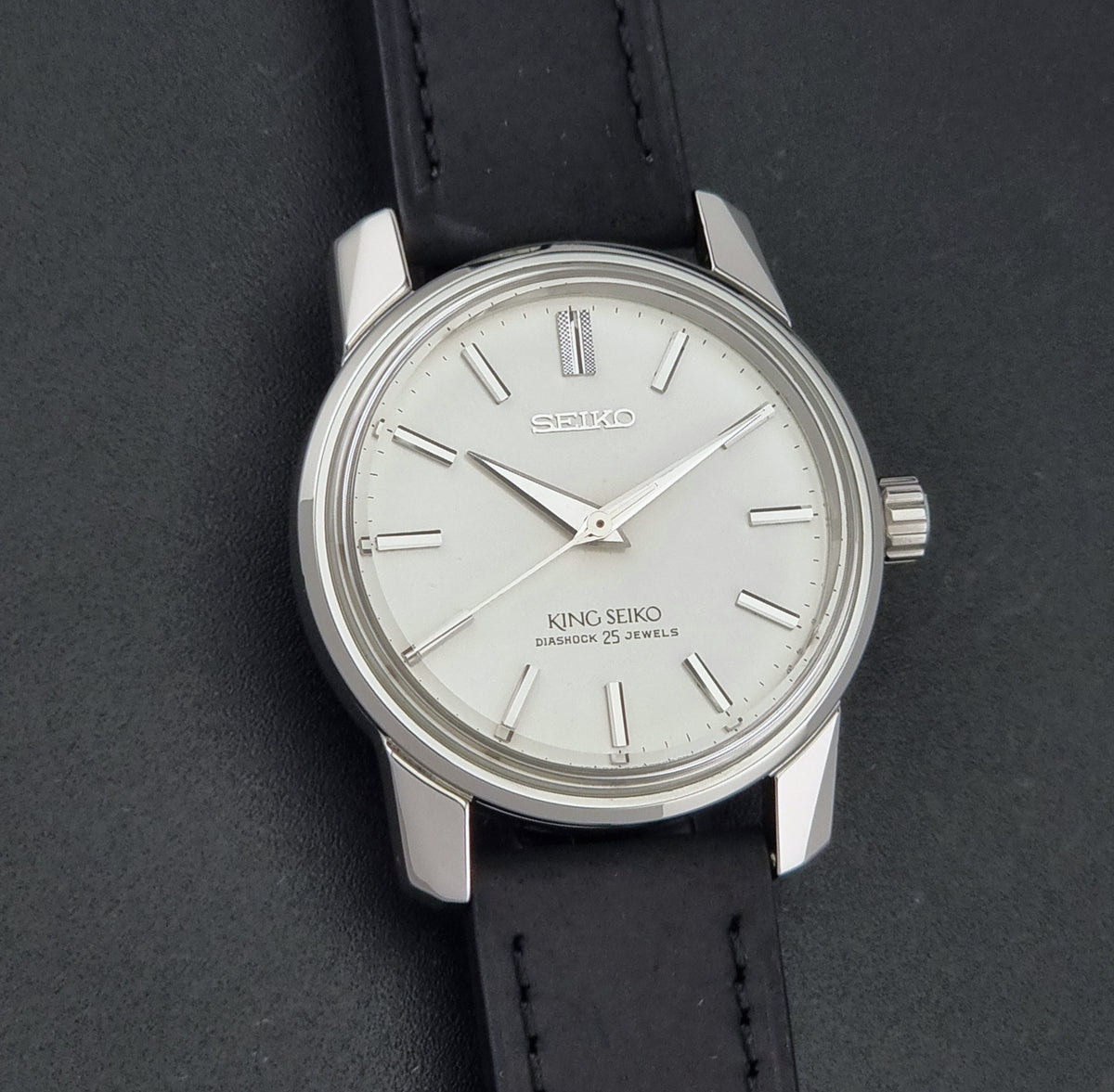 A vintage Seiko King Seiko watch with a silver-toned metal case, white dial, and black leather strap, shown on a dark textured background.