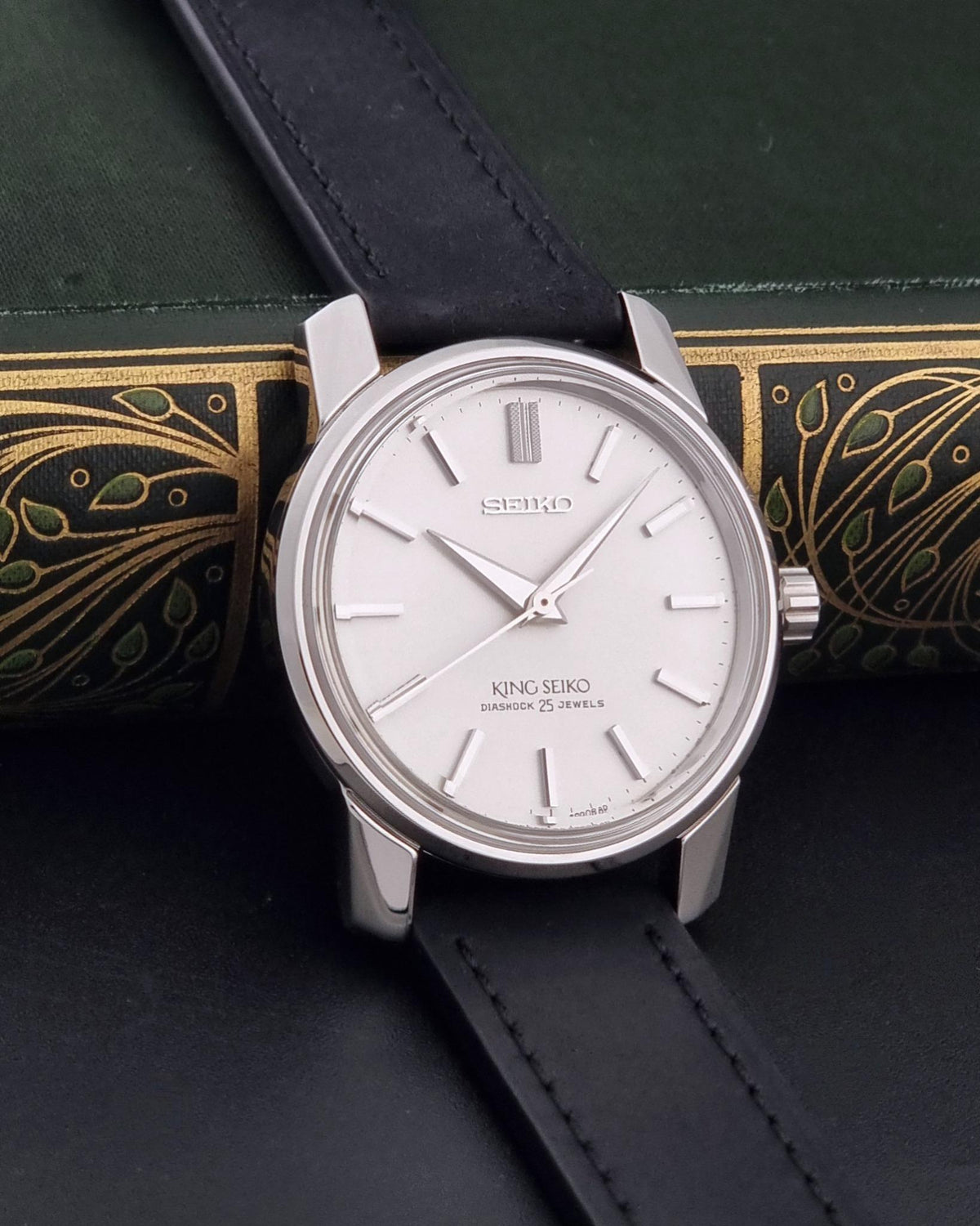 Vintage King Seiko watch 44-9990 with a silver-toned stainless steel case, white dial, and black leather strap, resting on a dark green book with gold detailing.