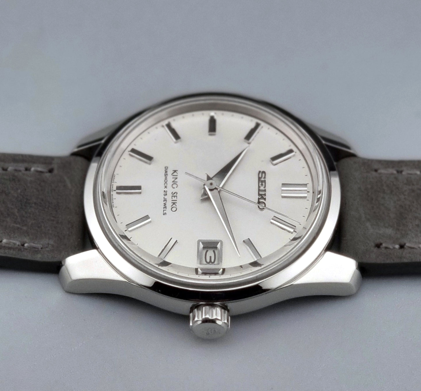 Vintage King Seiko 4402-8000 watch with a silver case and grey leather strap on a gray background angled view crown
