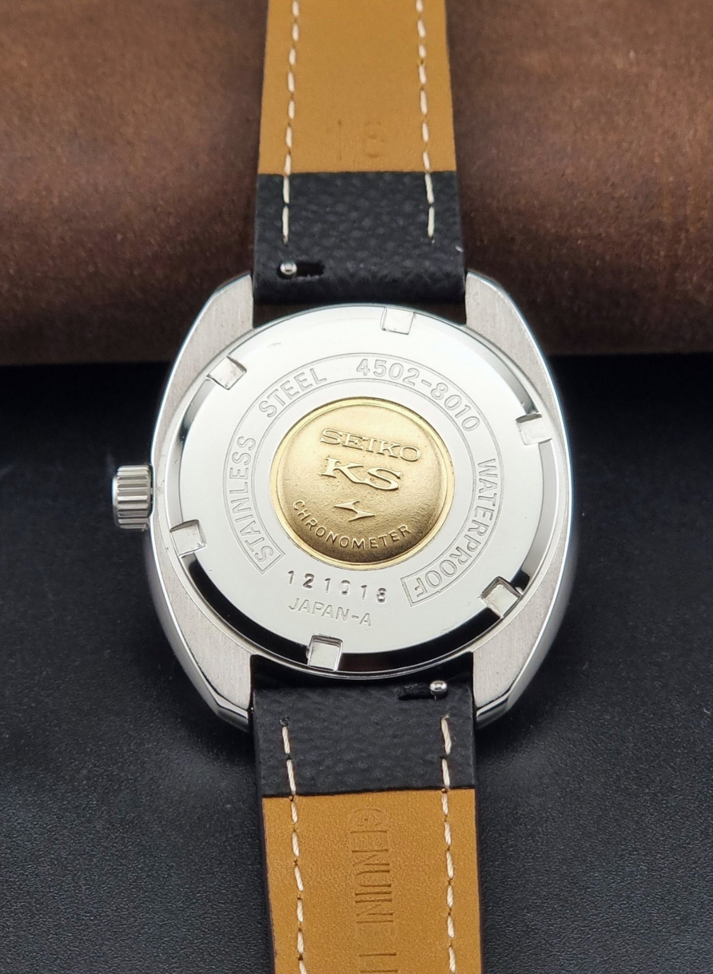 Vintage King Seiko Wristwatch 4502-8010 with a silver-toned metal case, white dial, and black textured leather strap, on a dark background case back view