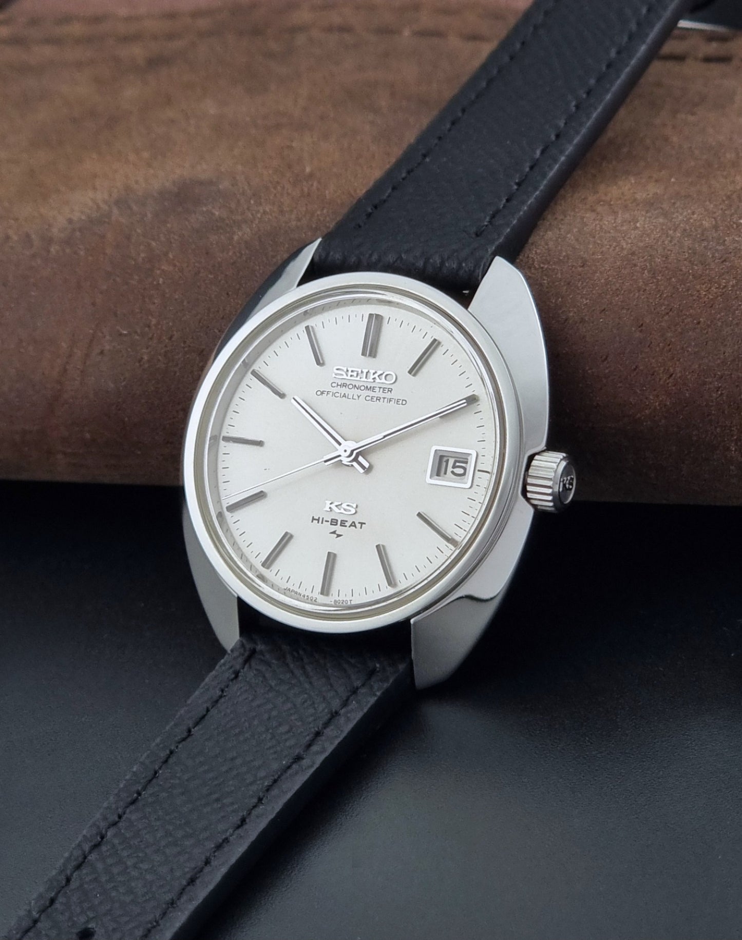 Vintage King Seiko Wristwatch 4502-8010 with a silver-toned metal case, white dial, and black textured leather strap, on a dark background crown view