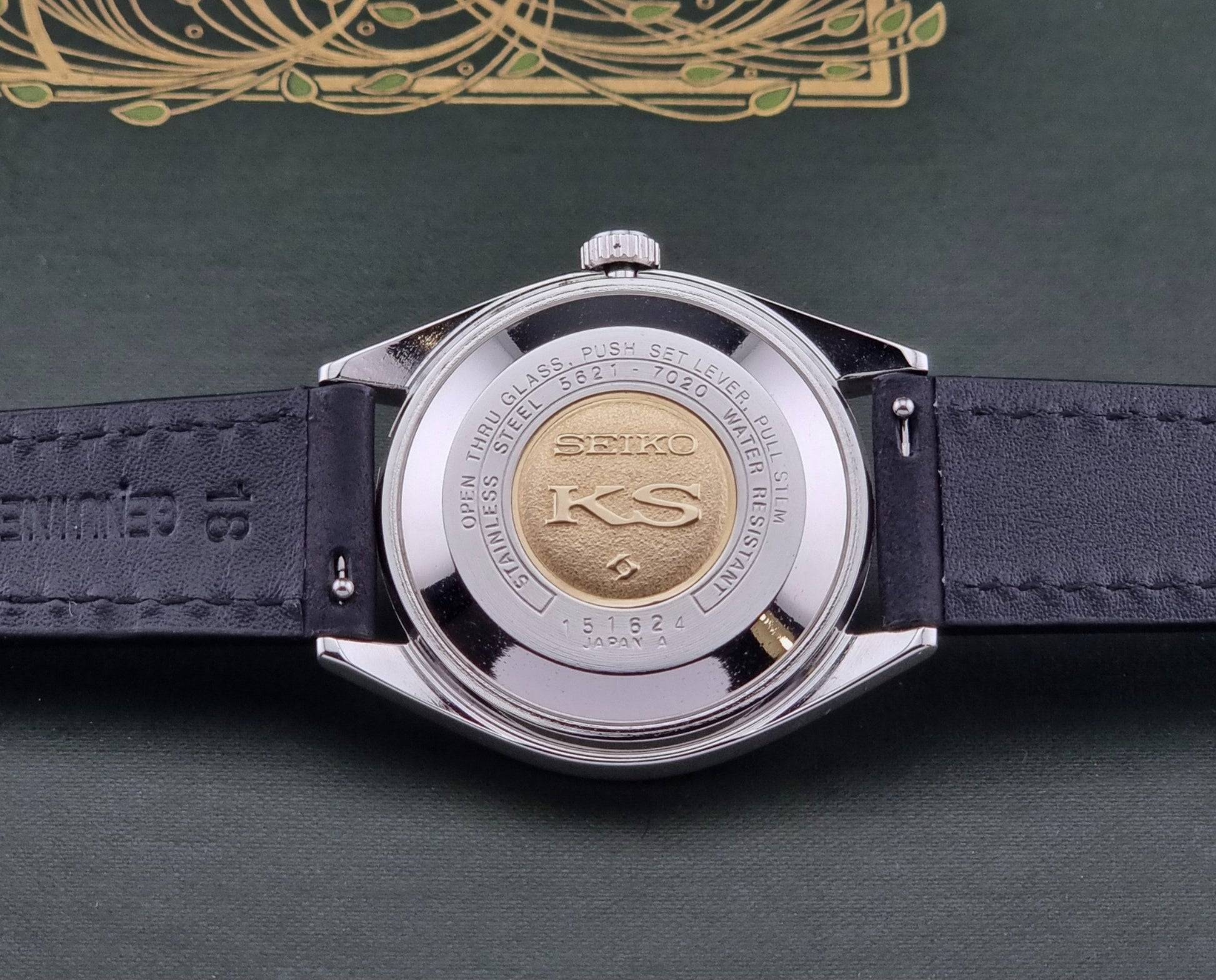 Close-up of a watch case back with a dark blue strap and silver case on a green background