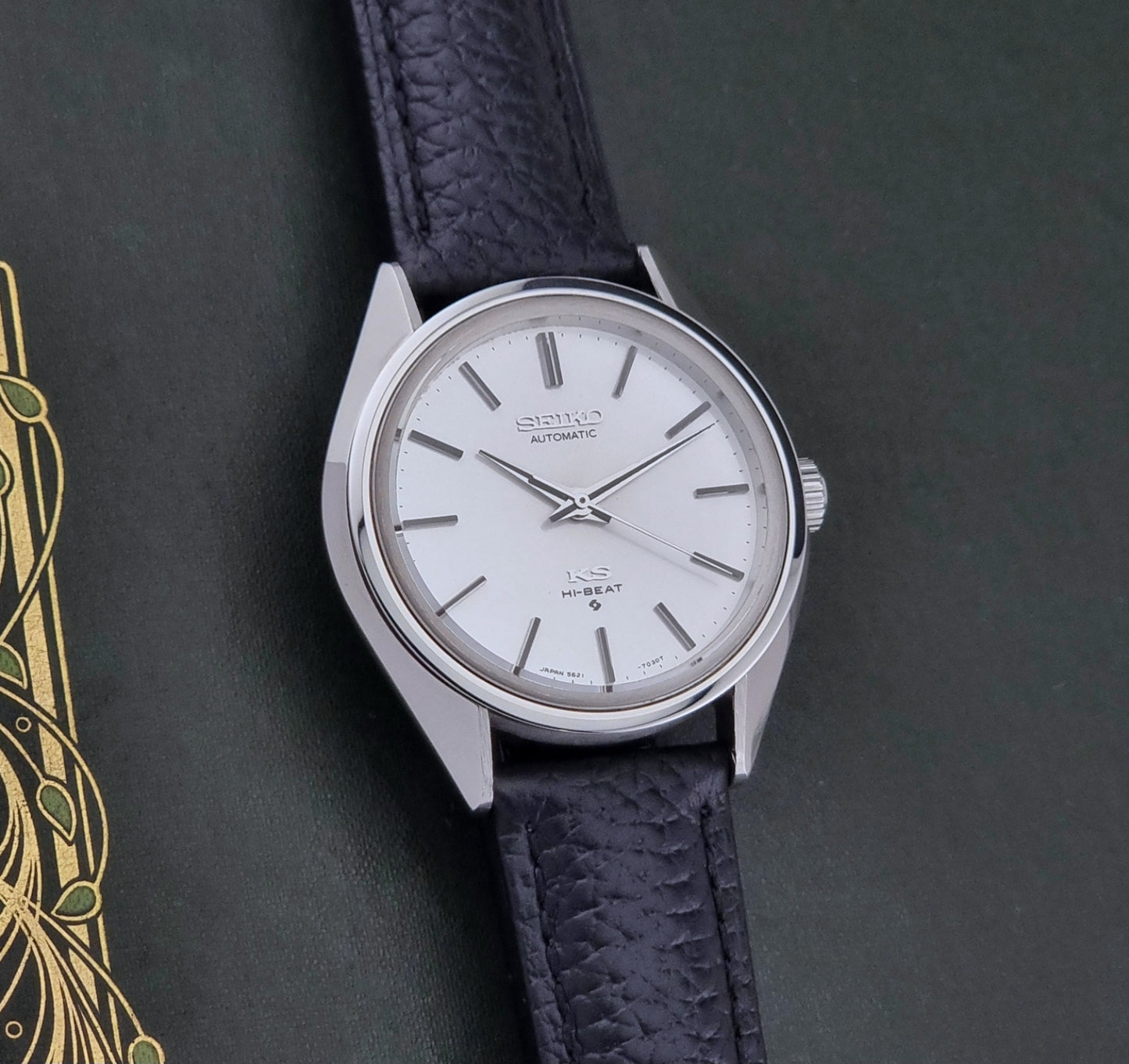 Vintage King Seiko Wristwatch 5621-7022 with a dark leather strap on a green background front view