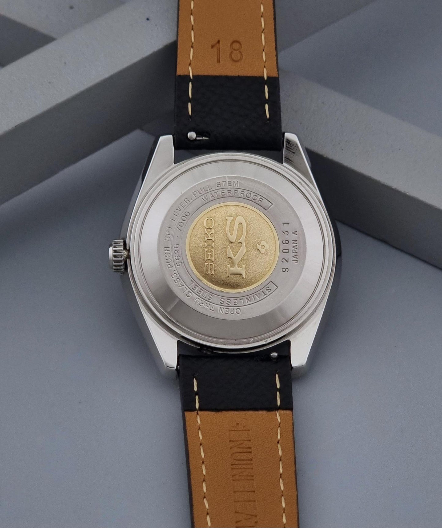 Close-up of the back of a Vintage King Seiko Wristwatch 5626-7000with a silver-toned case and a leather strap, resting on a gray geometric surface. The watch back shows a gold-toned circular emblem with the brand name visible.