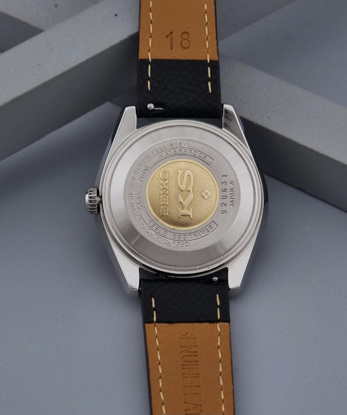 Close-up of the back of a Vintage King Seiko Wristwatch 5626-7000with a silver-toned case and a leather strap, resting on a gray geometric surface. The watch back shows a gold-toned circular emblem with the brand name visible.