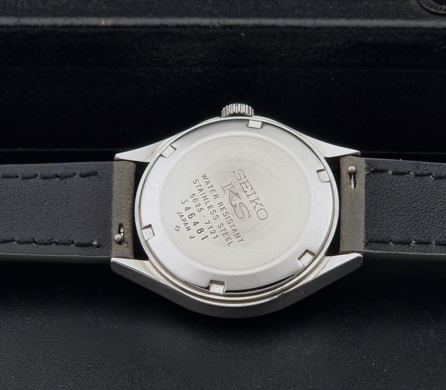 The back of a Vintage King Seiko 5625-7121 watch with a stainless steel case and a dark gray leather strap, resting on a black textured surface.