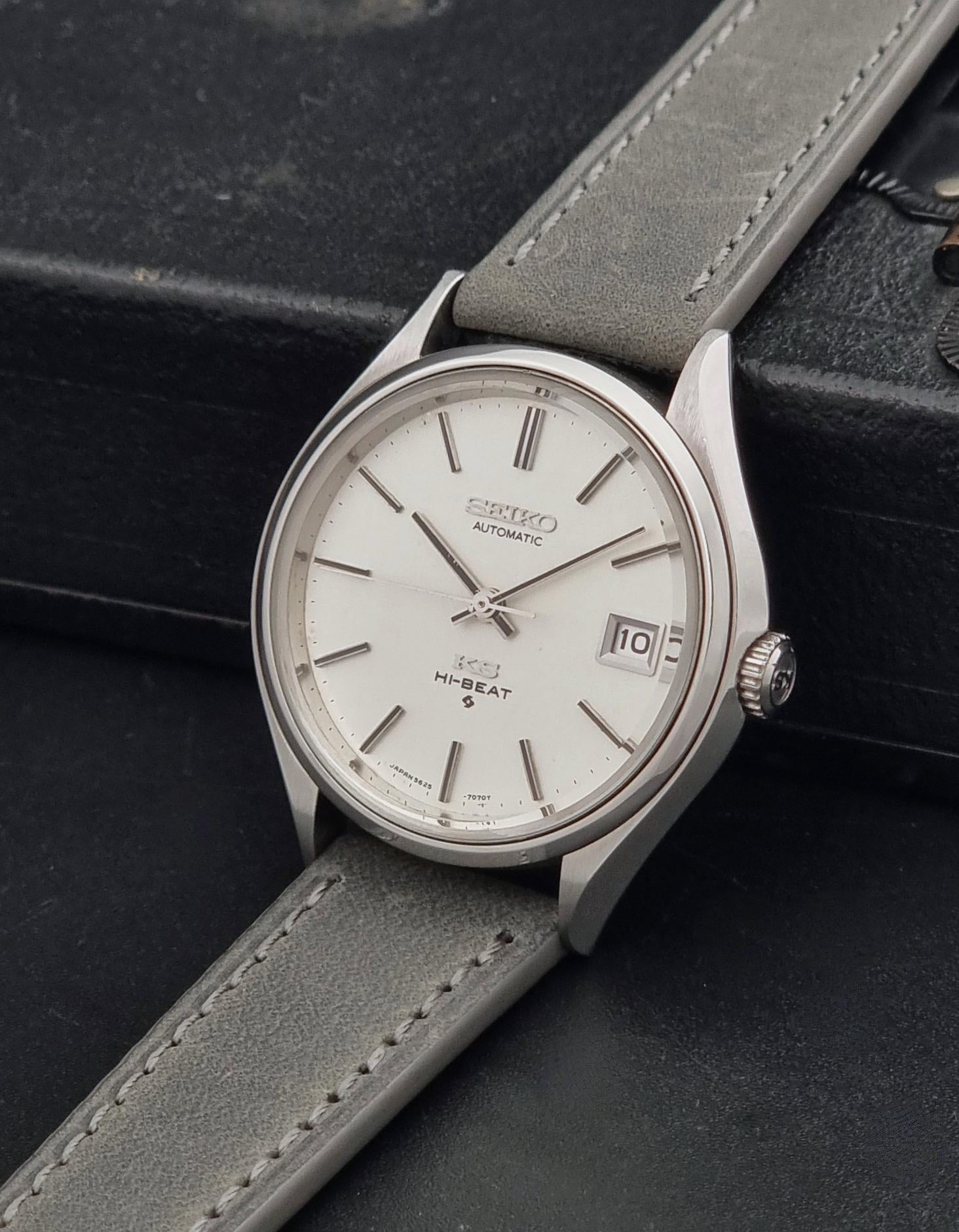 A Vintage King Seiko 5625-7121 watch with a silver-toned metal case, white dial, and a light gray leather strap, resting on a dark textured surface.