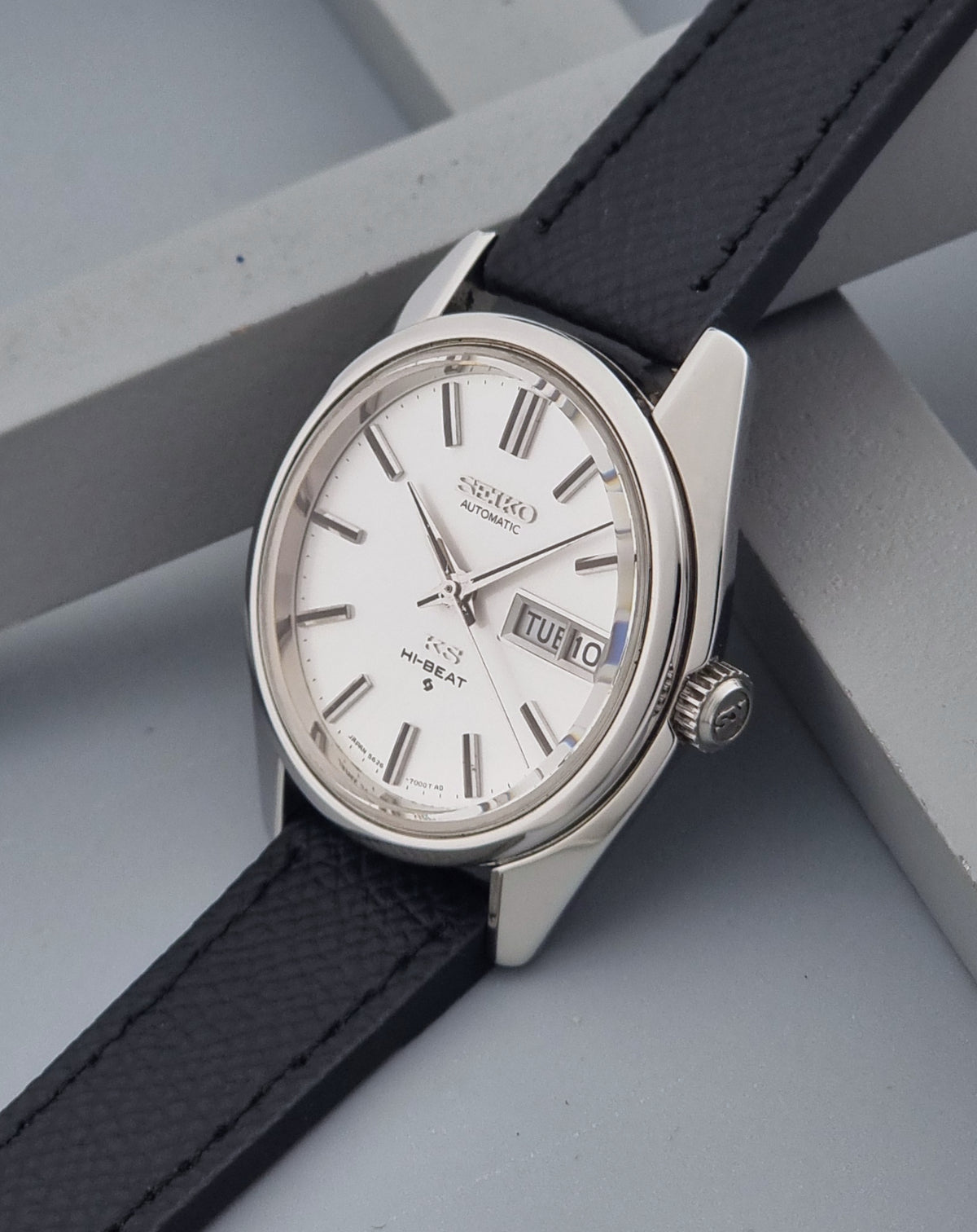 A Vintage King Seiko Wristwatch 5626-7000 with a silver-toned stainless steel case, white dial, and black textured leather strap, resting on a gray geometric background.