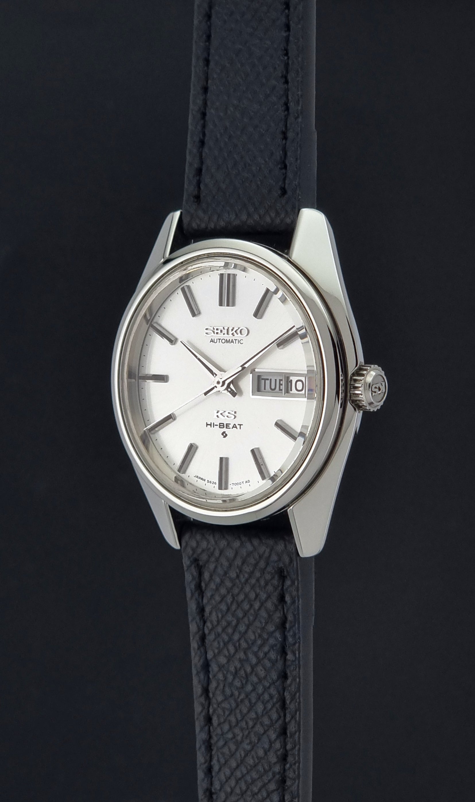 A Vintage King Seiko Wristwatch 5626-7000 with a silver-toned metal case, white dial, and textured dark strap, shown against a dark background.