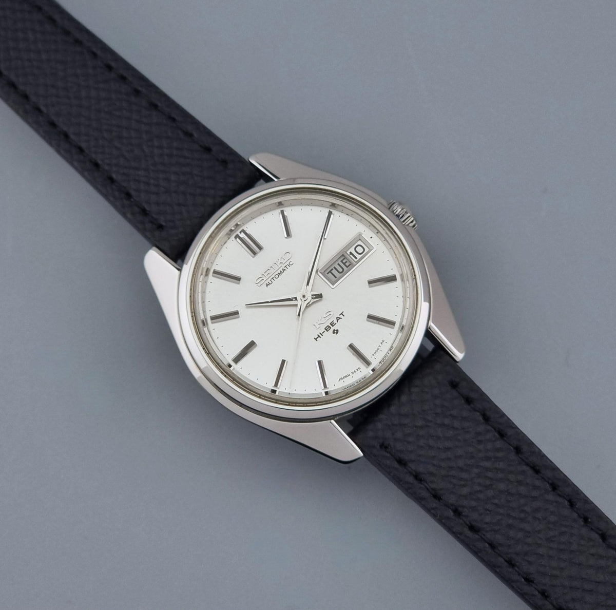 A Vintage King Seiko Wristwatch 5626-7000 with a silver-tone case, white dial, and textured black strap, photographed diagonally on a smooth gray background.