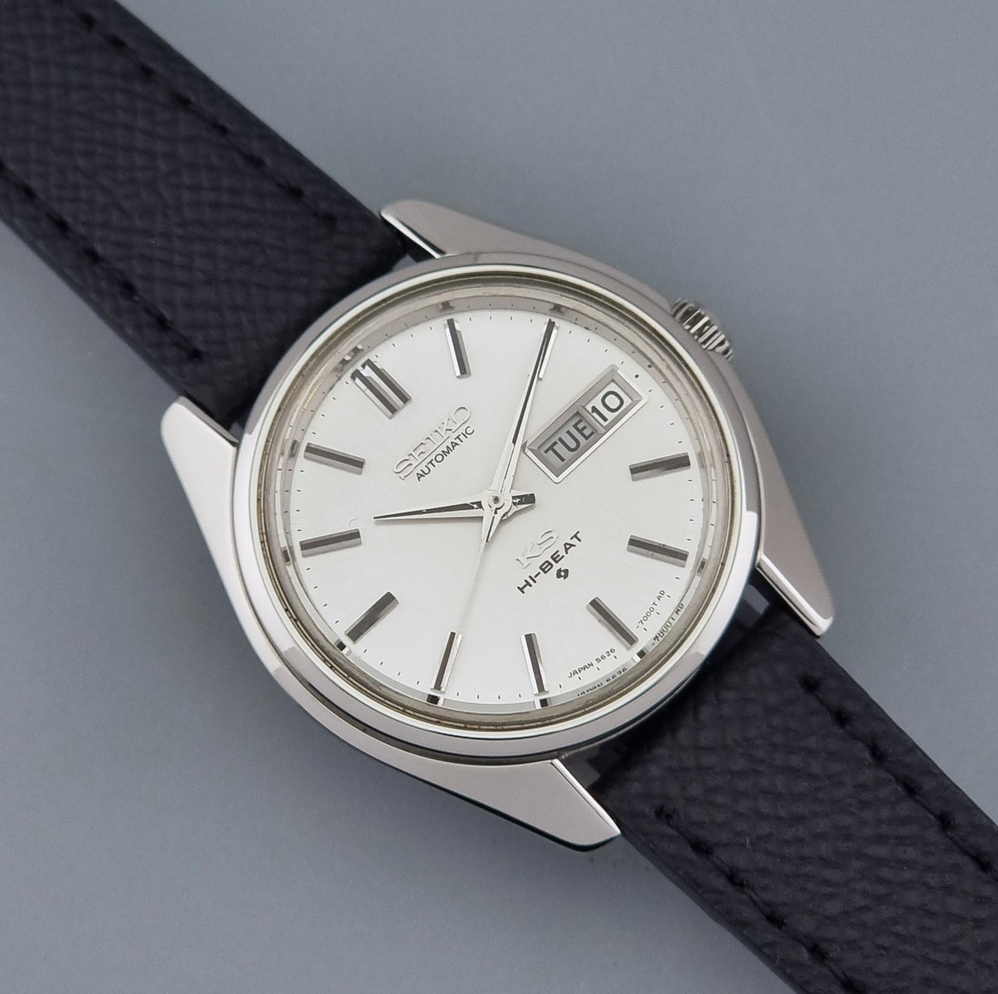 A Vintage King Seiko Wristwatch 5626-7000 with a silver stainless steel case, white dial, and a textured black leather strap, shown on a neutral gray background.