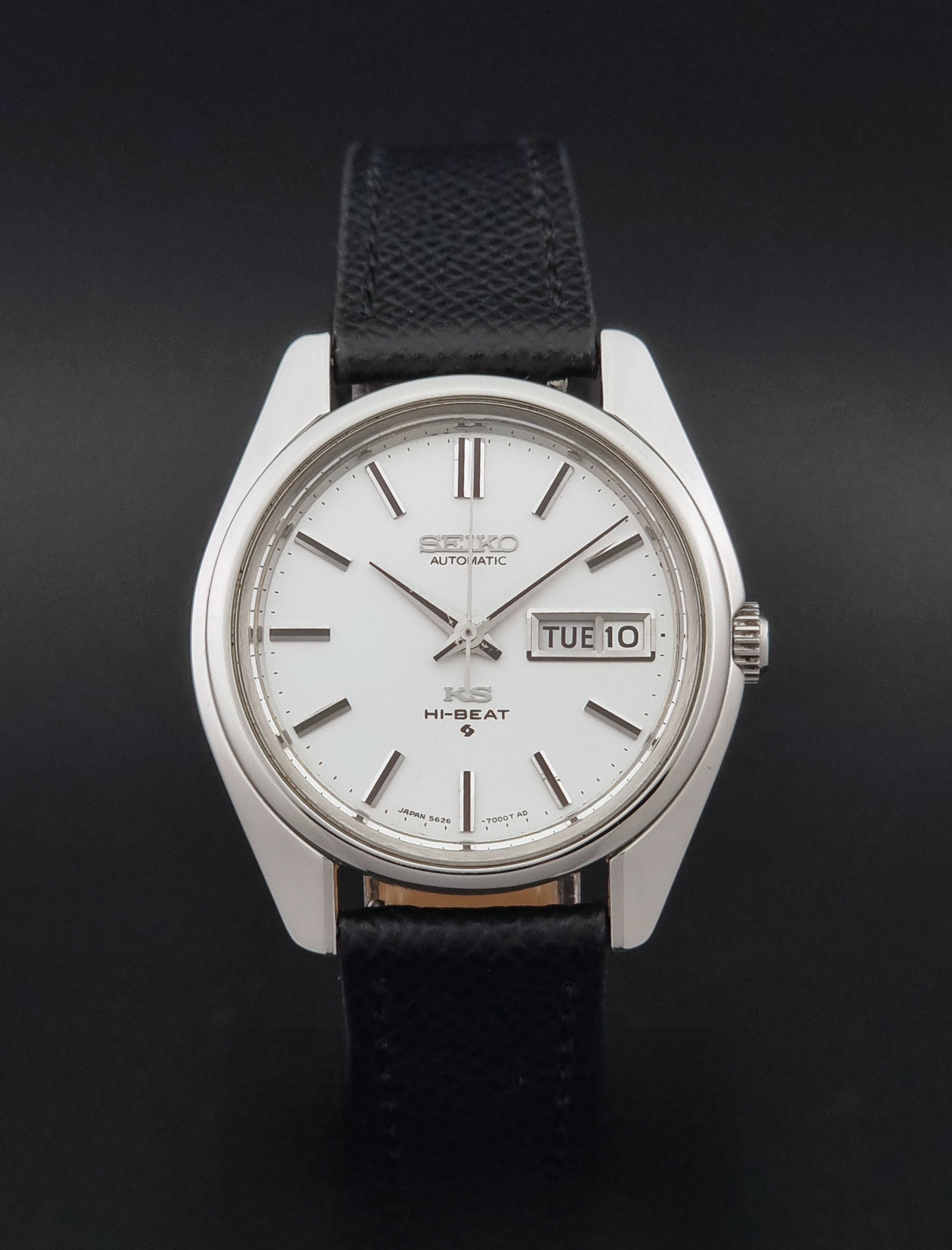 A Vintage King Seiko Wristwatch 5626-7000 with a silver-toned metal case, white dial, and black textured strap, photographed against a dark black background.