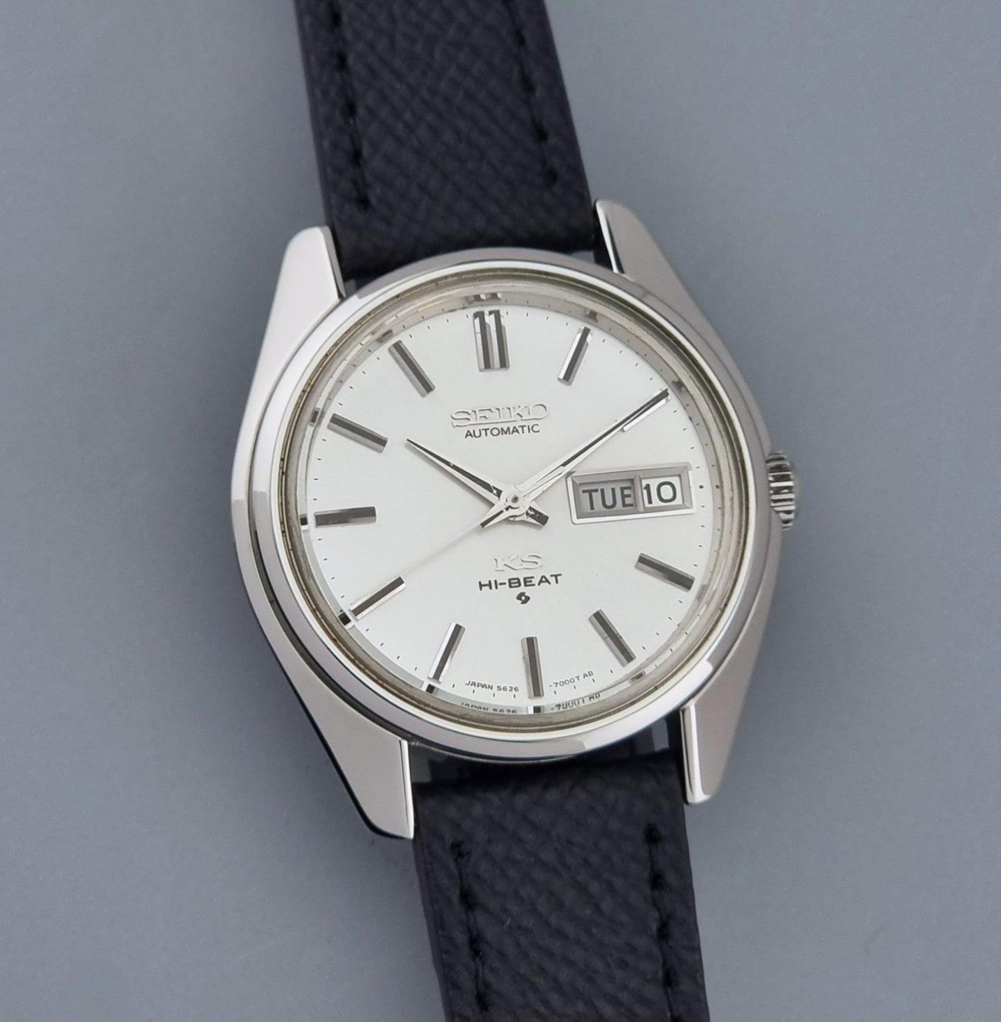 A Vintage King Seiko Wristwatch 5626-7000t automatic wristwatch with a silver-toned metal case, white dial, and a dark textured strap, shown against a solid gray background.