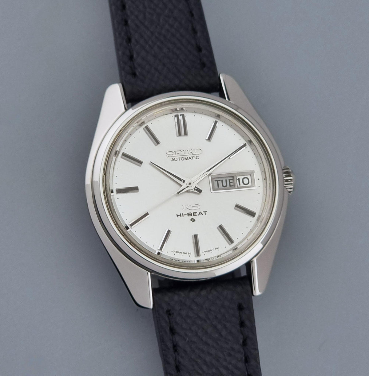 A Vintage King Seiko Wristwatch 5626-7000t automatic wristwatch with a silver-toned metal case, white dial, and a dark textured strap, shown against a solid gray background.