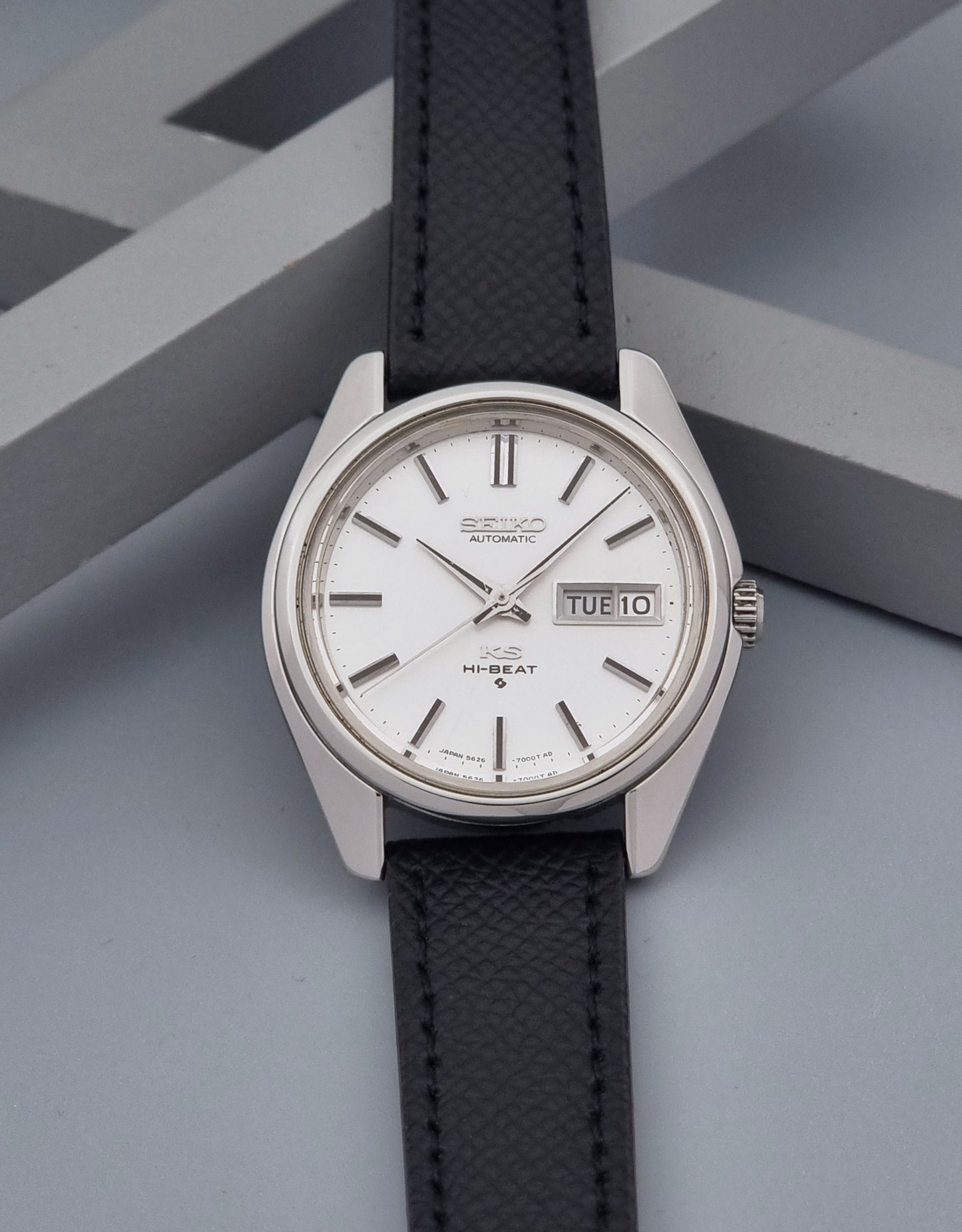 A Vintage King Seiko Wristwatch 5626-7000 Hi-Beat wristwatch with a silver-toned metal case, white dial, and black textured leather strap, displayed on a gray geometric background.