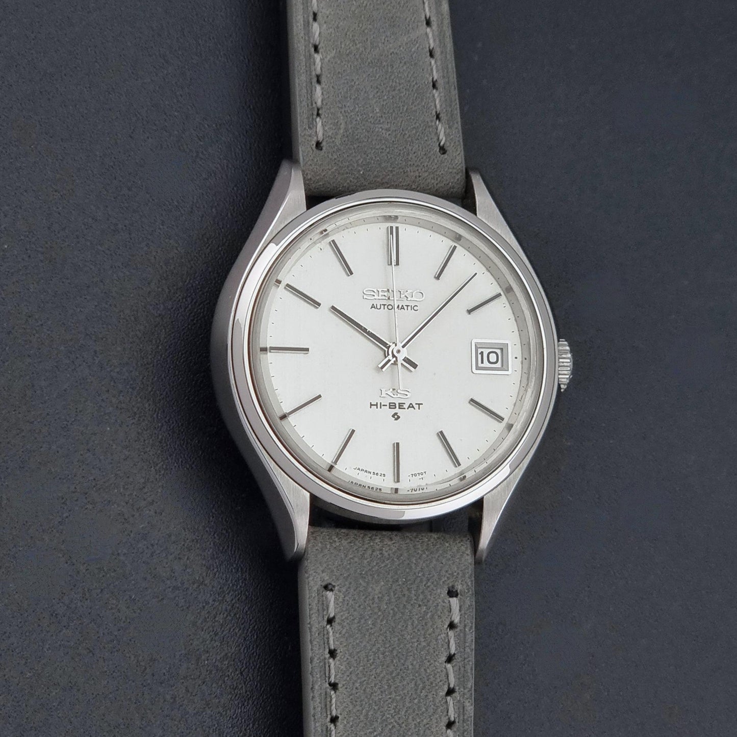 A Vintage King Seiko 5625-7121 watch with a silver-toned metal case, white dial, and a gray leather strap, shown on a dark textured background.