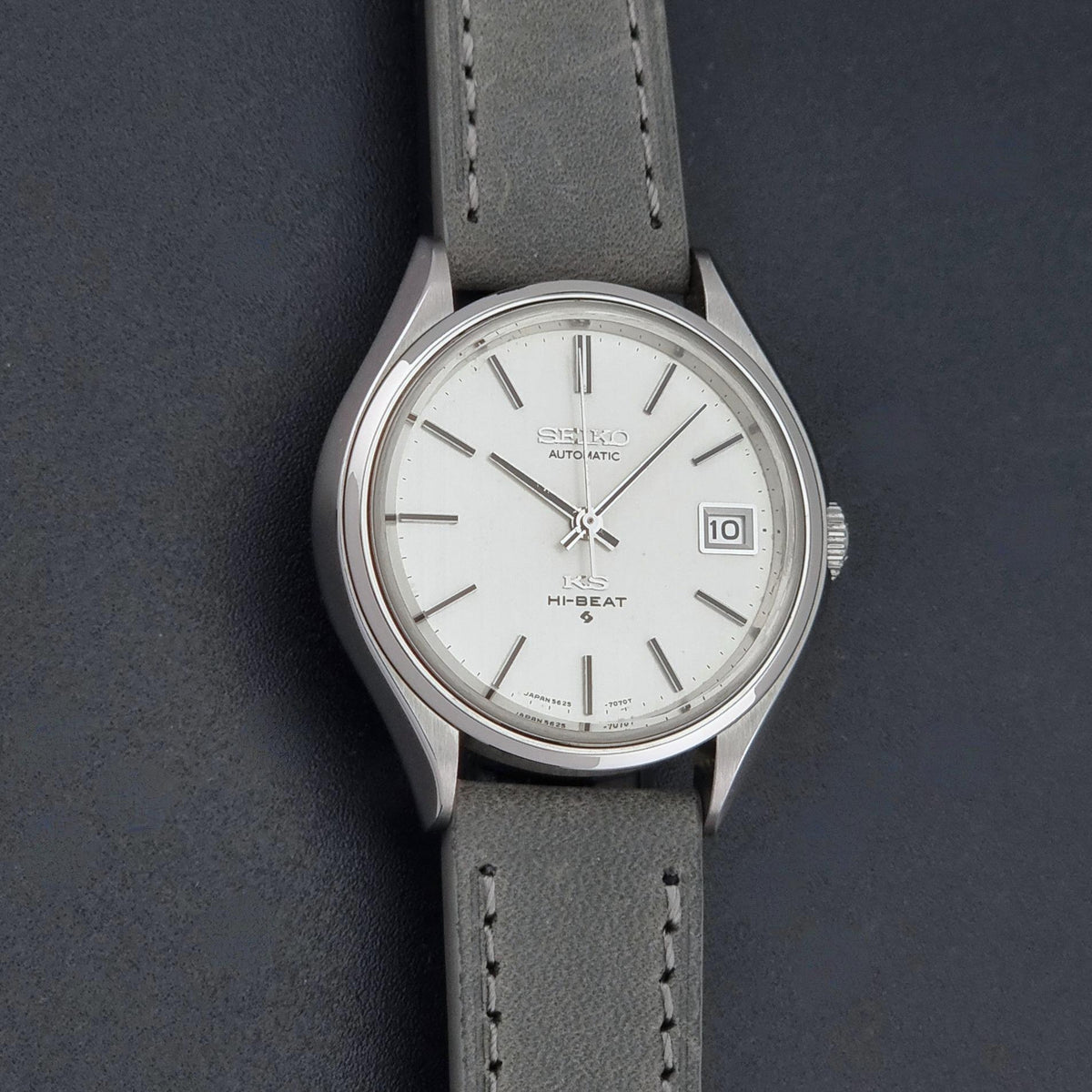 A Vintage King Seiko 5625-7121 watch with a silver-toned metal case, white dial, and a gray leather strap, shown on a dark textured background.