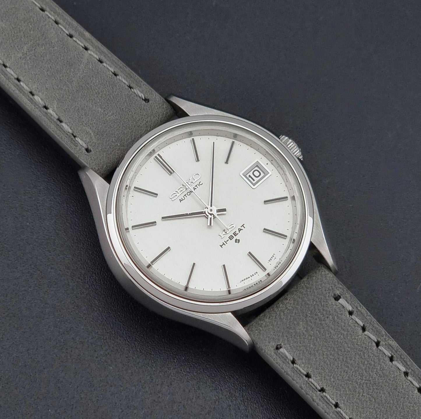 A Vintage King Seiko 5625-7121 watch with a white dial, silver-tone case, and a gray leather strap, shown on a dark textured background.