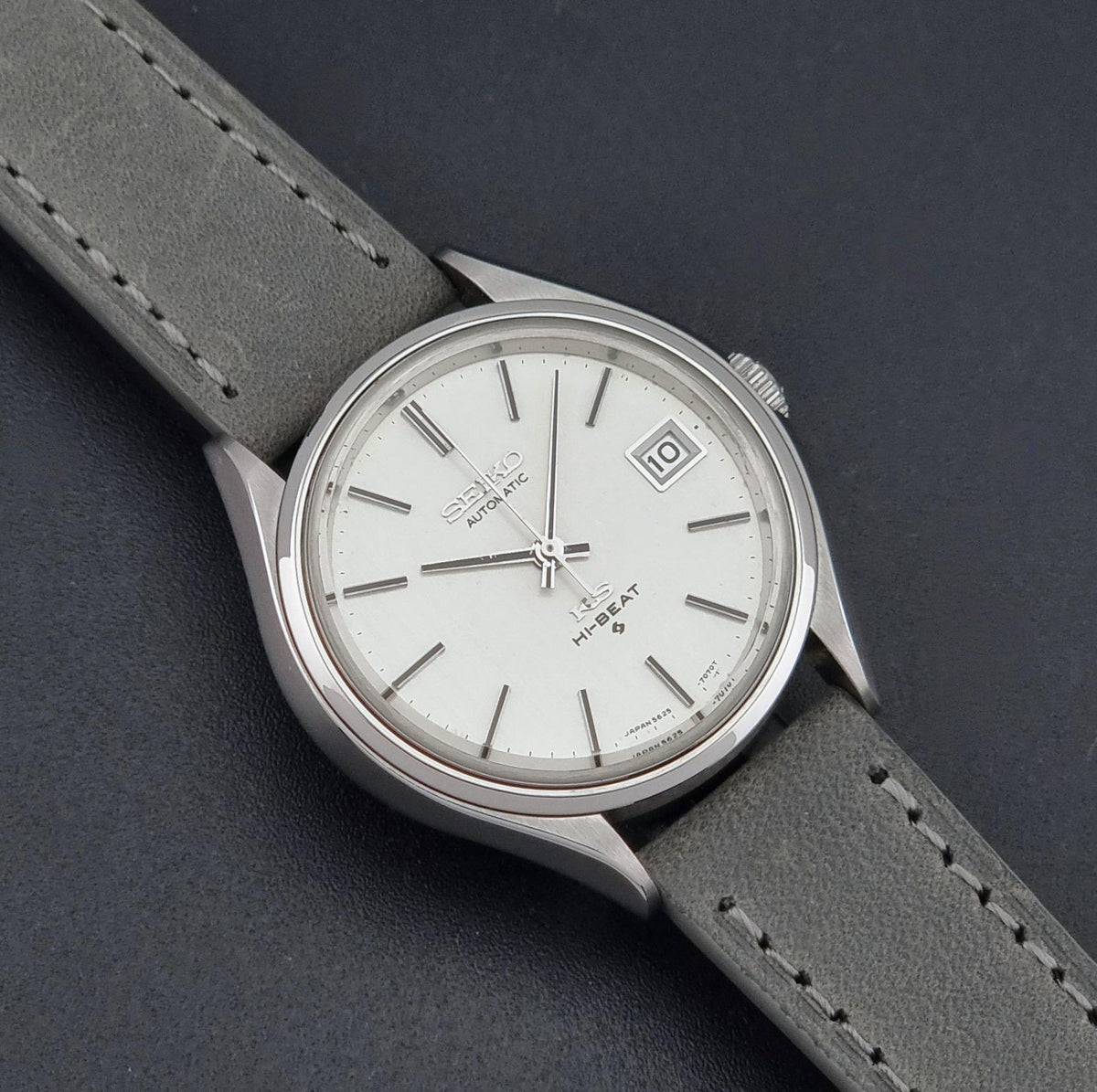 A Vintage King Seiko 5625-7121 watch with a white dial, silver-tone case, and a gray leather strap, shown on a dark textured background.